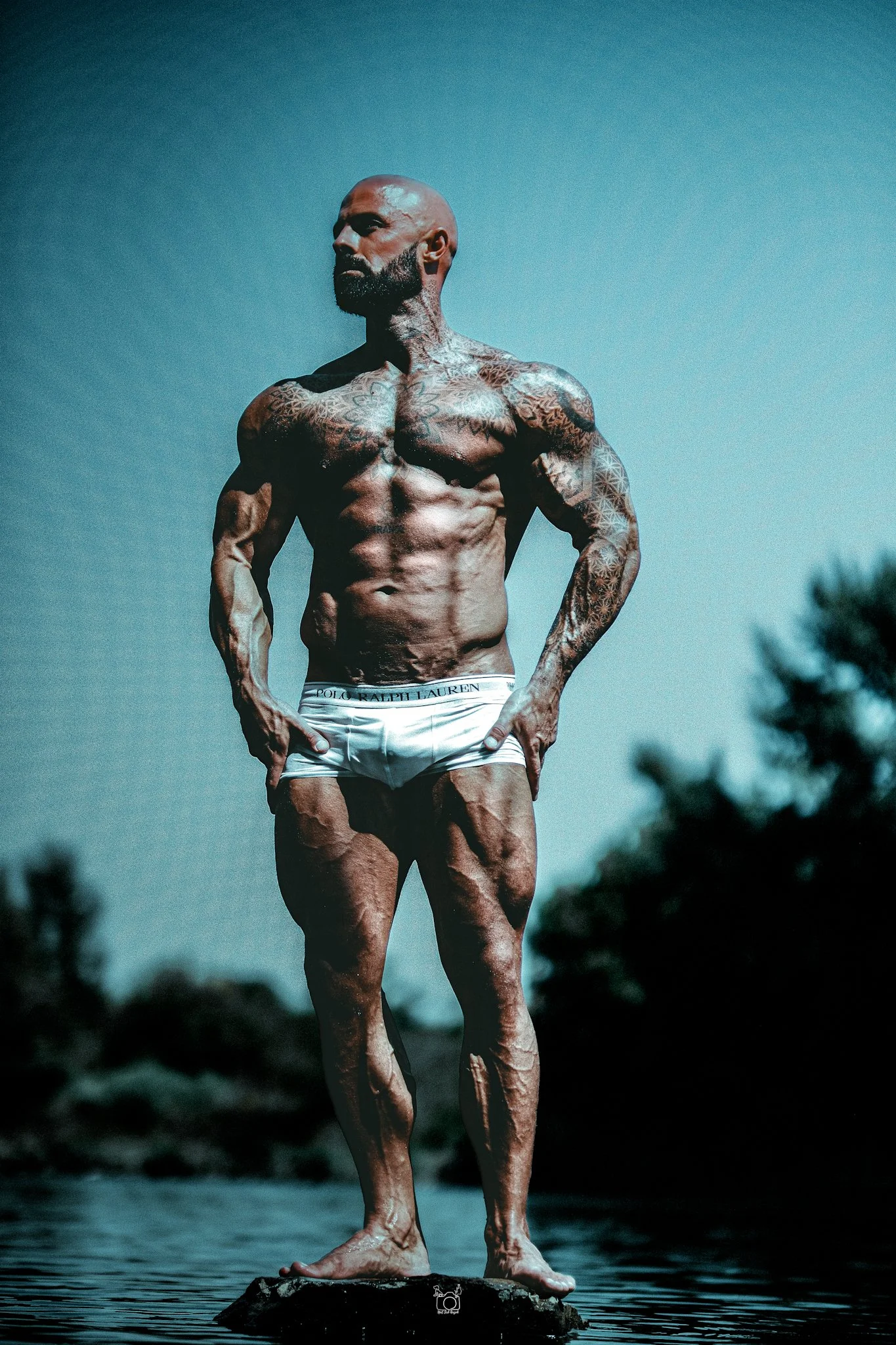 A muscular man with tattoos standing on a rock in water, wearing white underwear, with a clear sky in the background.