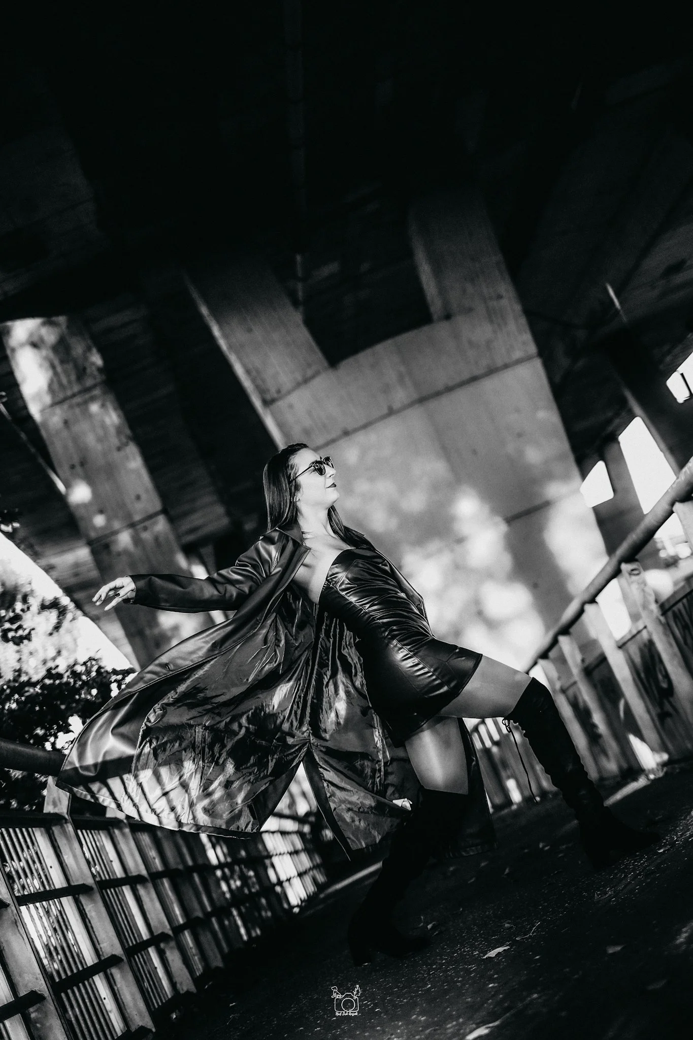 A woman in sunglasses and a shiny trench coat poses under a bridge, with her arm outstretched and her back arched, in an urban setting, black and white photo.