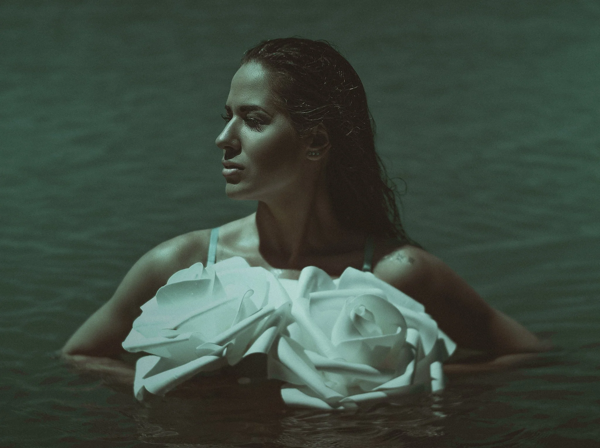 A woman with wet hair and closed eyes, wearing a white dress with ruffles, stands in water with a neutral expression.