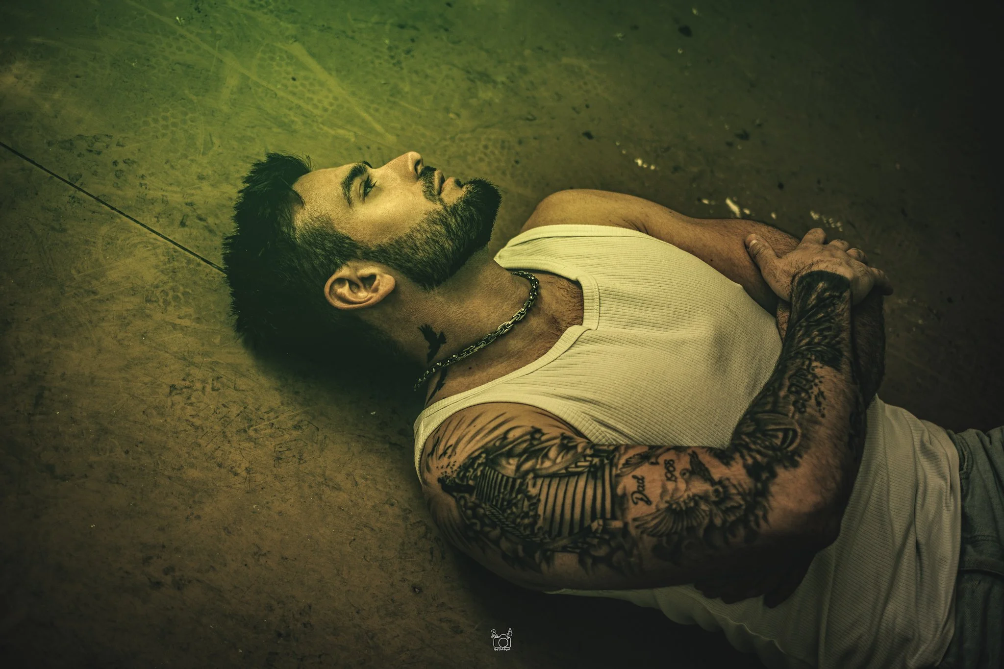 A man with tattoos and a beard lying on the ground with his eyes closed. He is wearing a white tank top and a chain necklace. Someone is holding his hand.