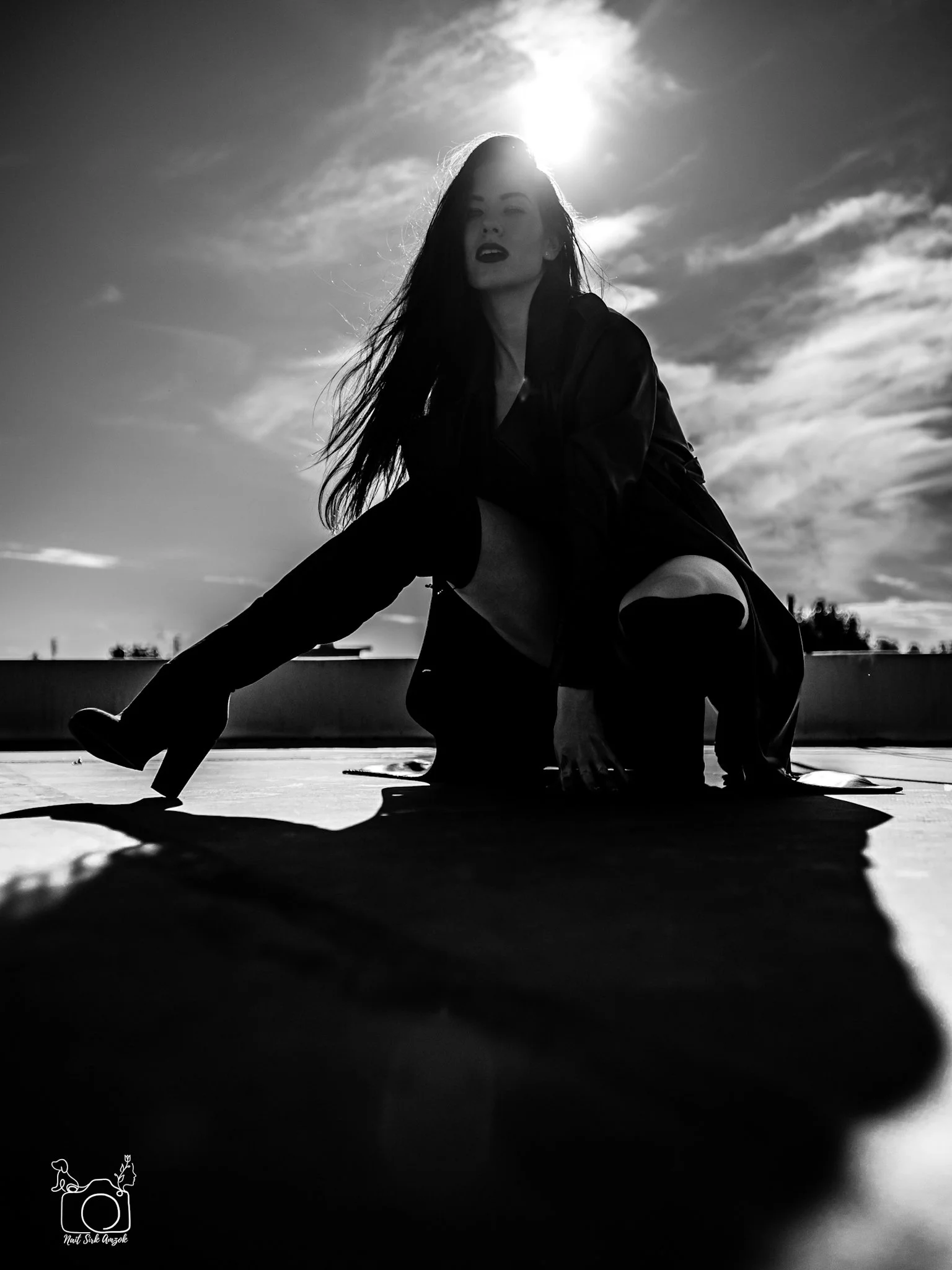 Black and white photo of a woman with long hair, sitting outdoors with the sun shining behind her. She is looking directly at the camera with a serious expression.