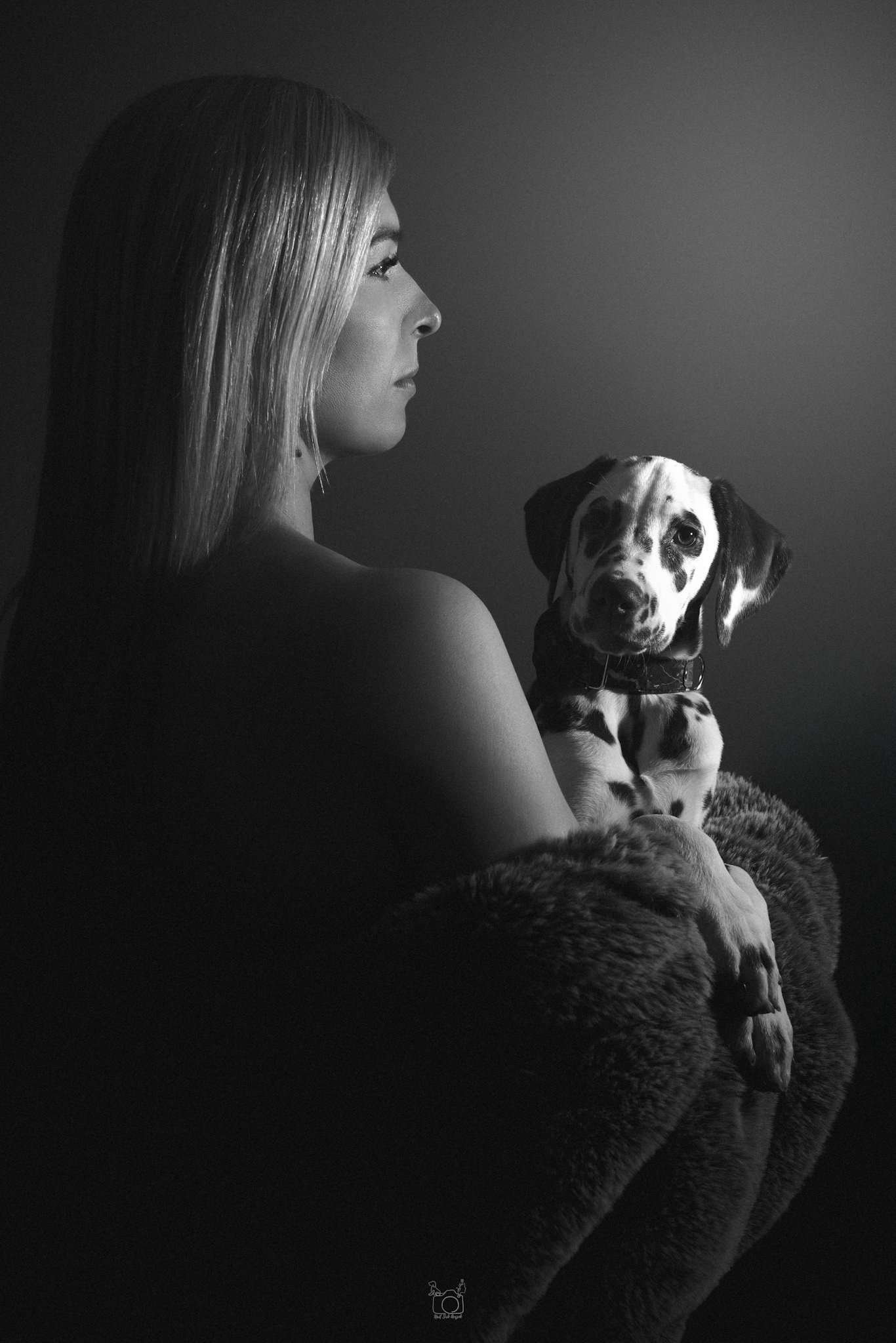 A black-and-white photo of a woman and a Dalmatian dog sitting together, both facing left. The woman has straight hair and a serious expression, while the dog has distinctive black spots and a calm look.