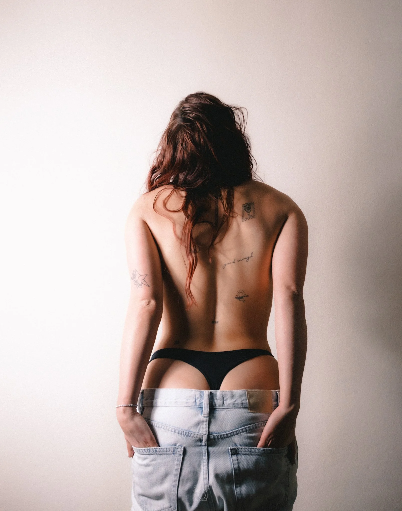 Woman with long reddish-brown wavy hair in black underwear and blue jeans, showing tattoos on her back and arms, standing with back to the camera against a plain white wall.
