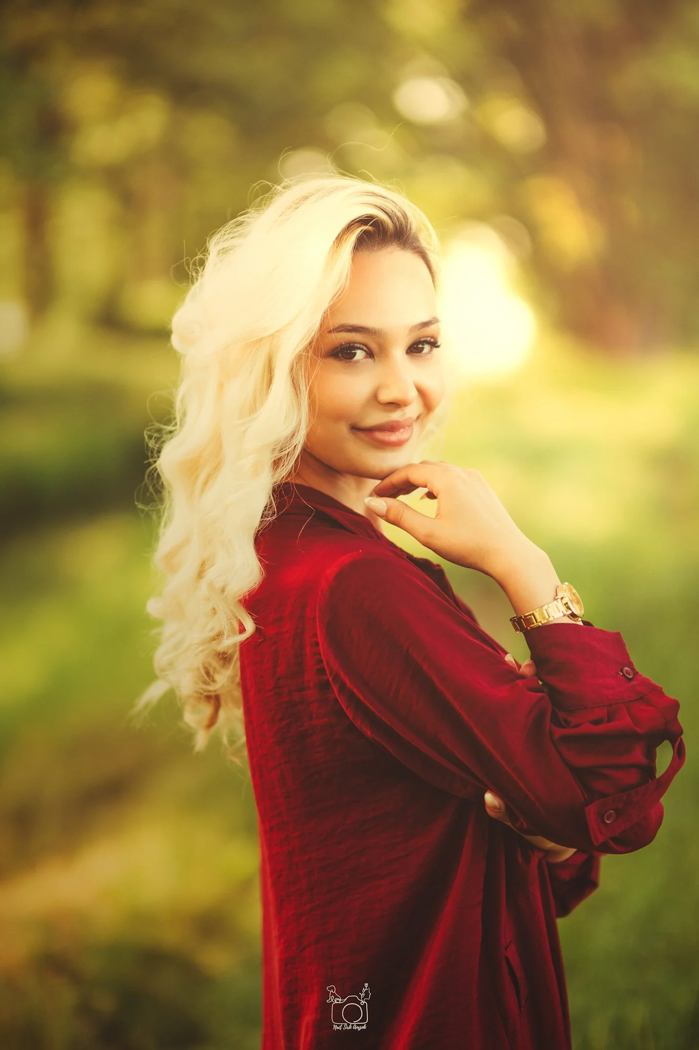 A woman with long blonde hair wearing a red velvet dress and gold watch, standing outdoors in a natural setting with blurred trees and sunlight in the background.