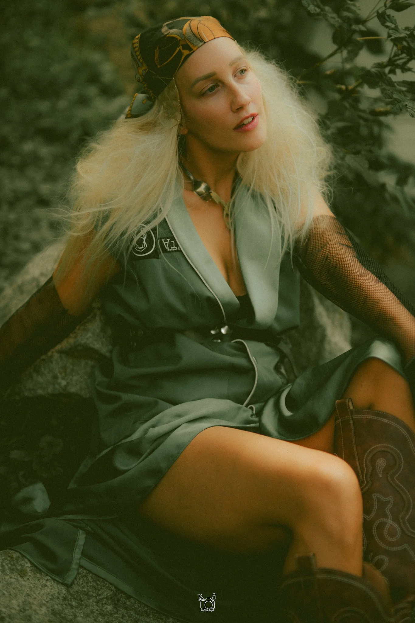 A woman with long blond hair and light skin is sitting outside on the ground, leaning back against a large rock or concrete surface. She is wearing a patterned headscarf, a gray vest with logos, mesh sleeves, shorts, and boots. She appears to be look
