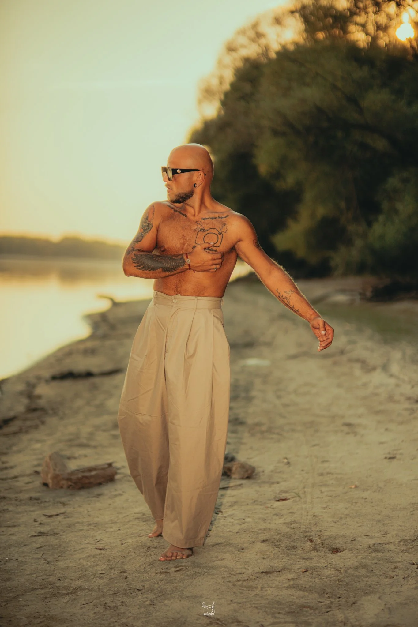 A shirtless man with tattoos and sunglasses walking barefoot on a sandy beach at sunset, with trees and the water in the background.