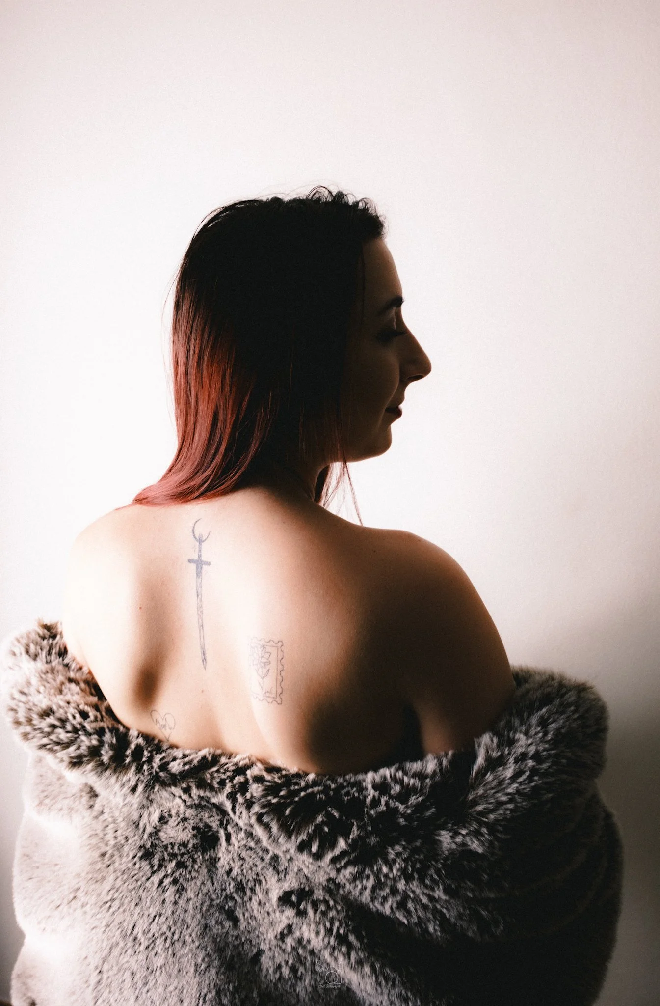 A woman with long dark hair, wearing a faux fur coat, is seen from the back with her face in profile. She has tattoos on her upper back and shoulder, including a sword, a stamp, and smaller designs.