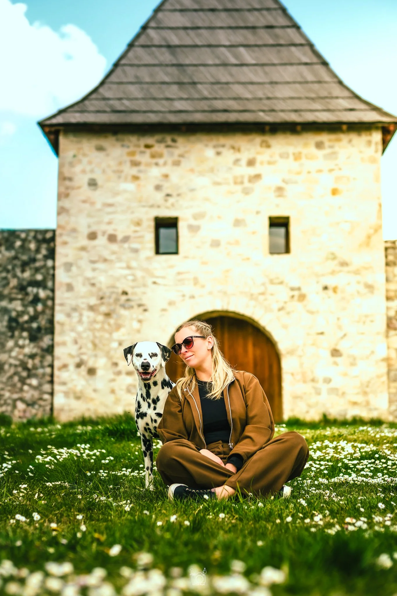 A woman in sunglasses sitting on grass with small white flowers, next to a Dalmatian dog, in front of an old stone building with a wooden door.