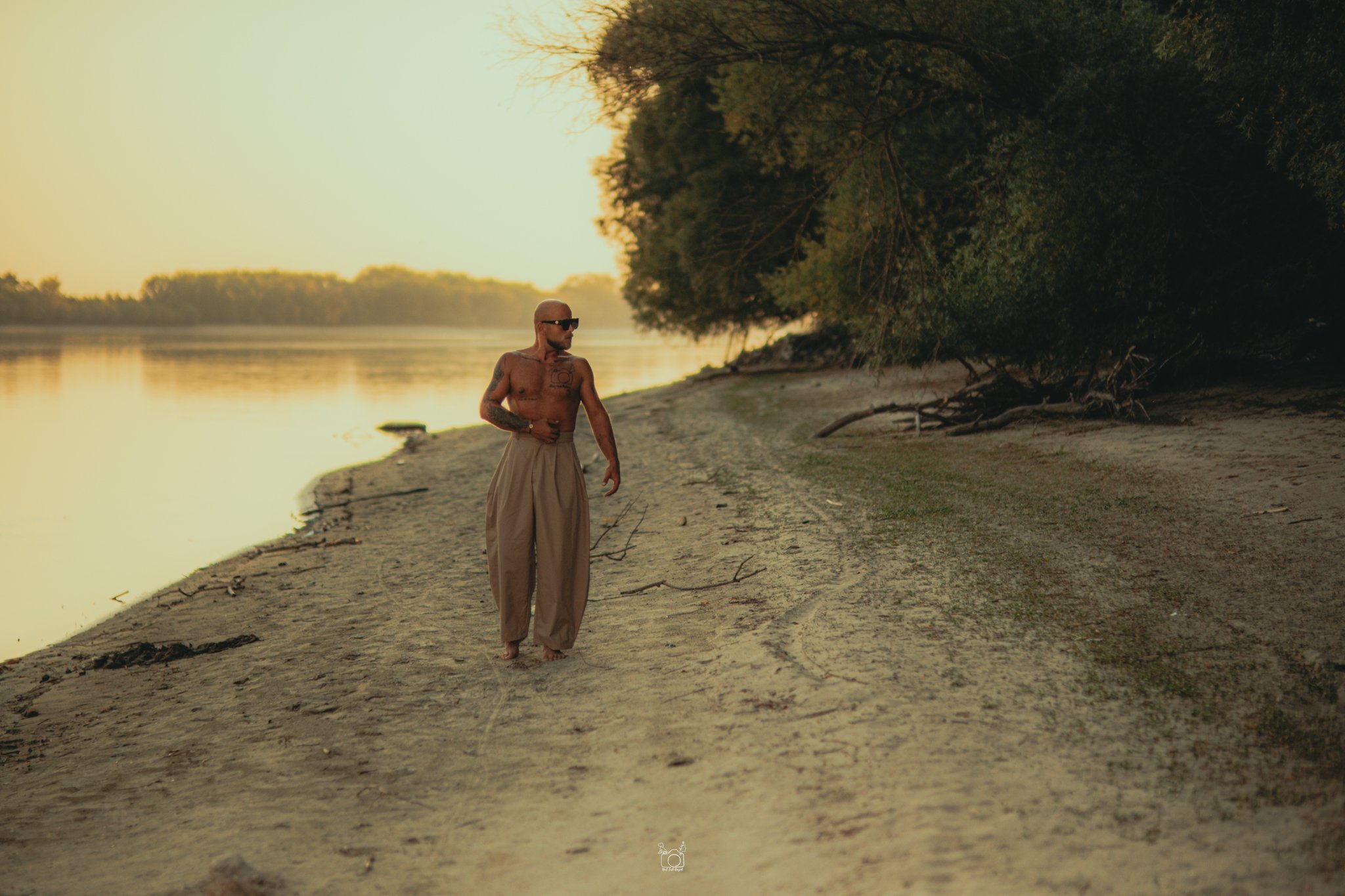 A shirtless man with tattoos and sunglasses walking on a sandy riverbank at sunset, with trees and water in the background.