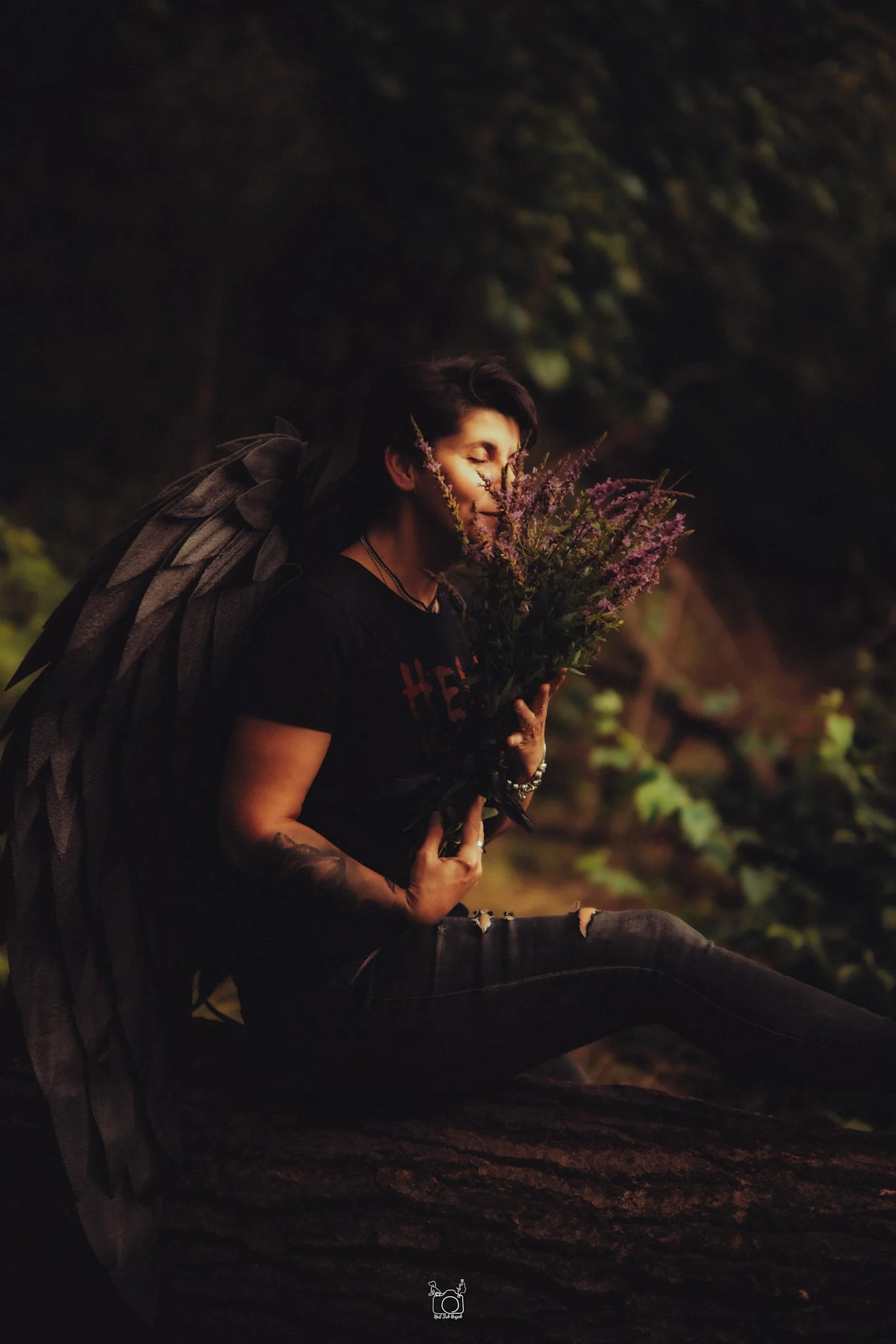 A person with short dark hair and tattoos on their arm, dressed in black and wearing jewelry, is sitting outdoors at night, holding a bouquet of purple flowers close to their face with eyes closed, smiling gently, with trees and dark foliage in the b