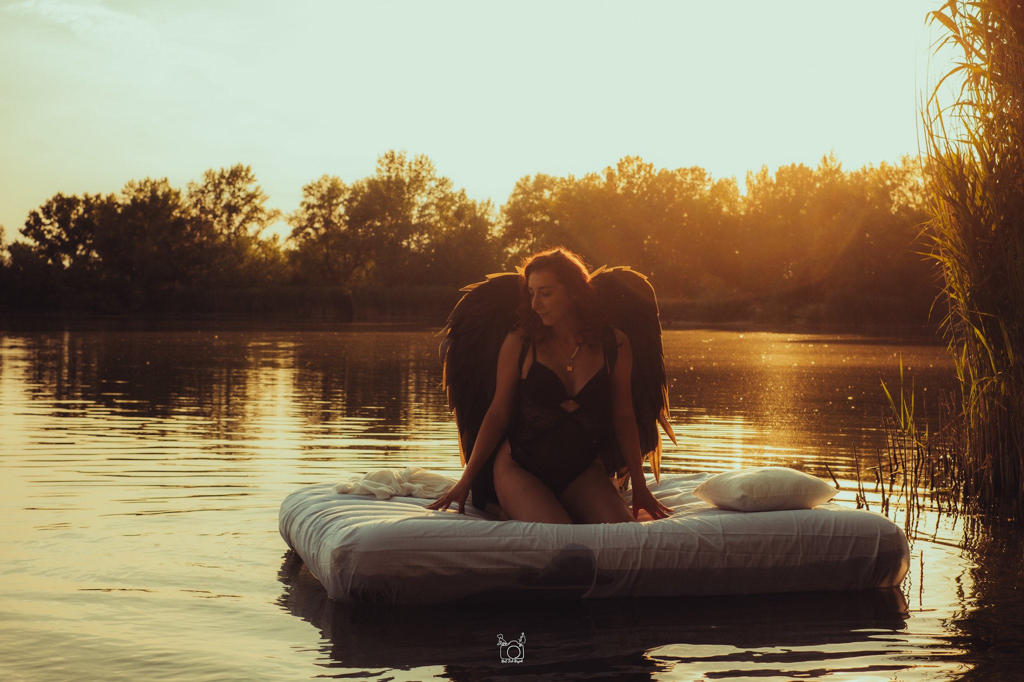 A woman with angel wings sitting on a floating platform in a river at sunset, with trees and tall grass in the background.