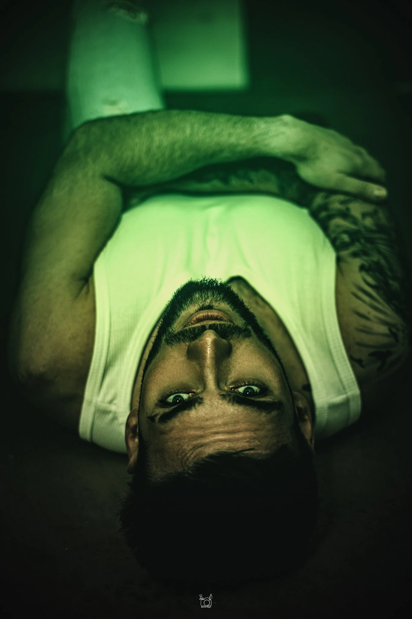 A man with glasses and a beard lying on the floor, looking directly at the camera, with his head upside down and his hand resting on his arm.