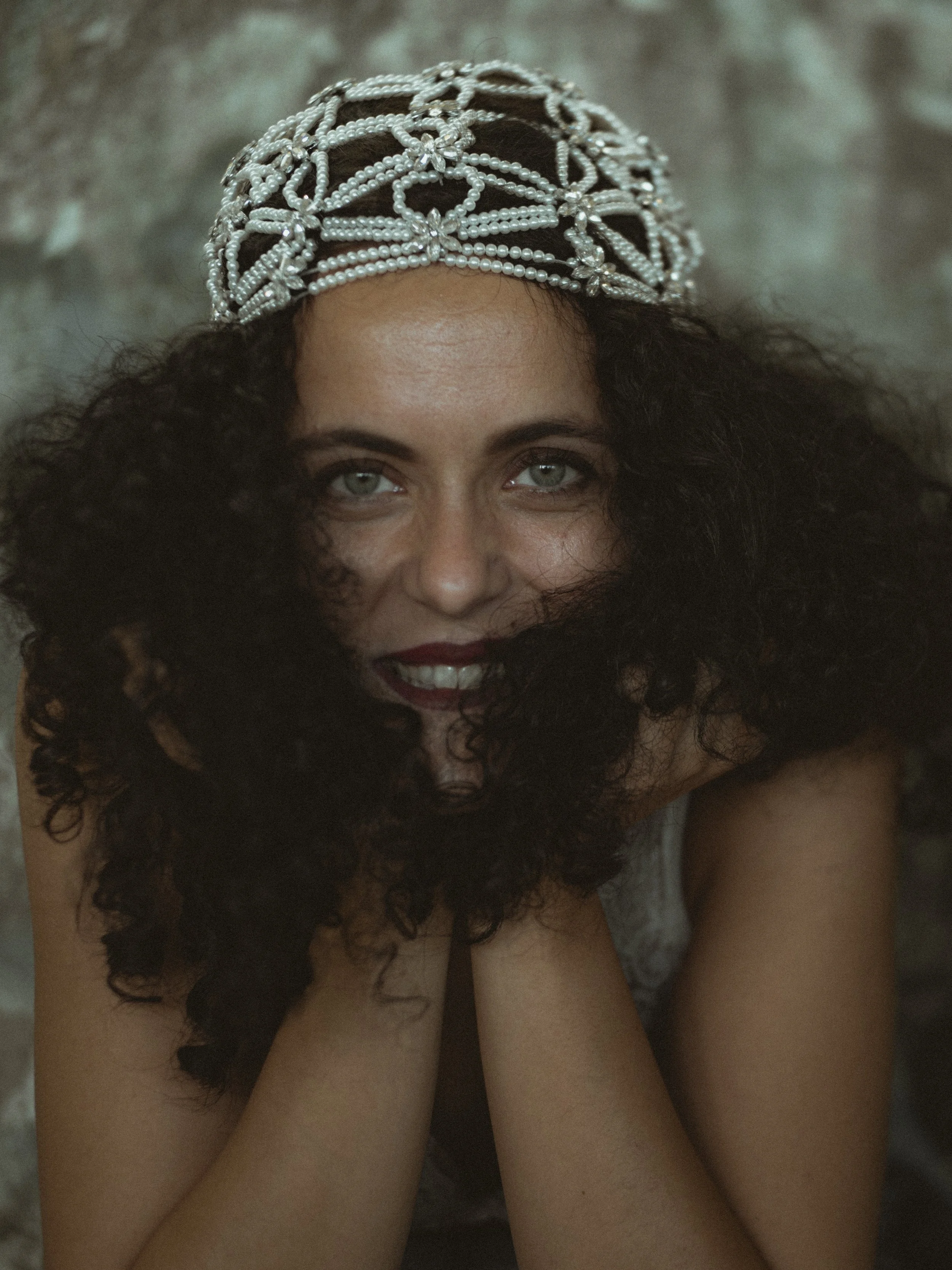 A woman with curly black hair wears a pearl and rhinestone beaded headpiece and has emerald green eyes.