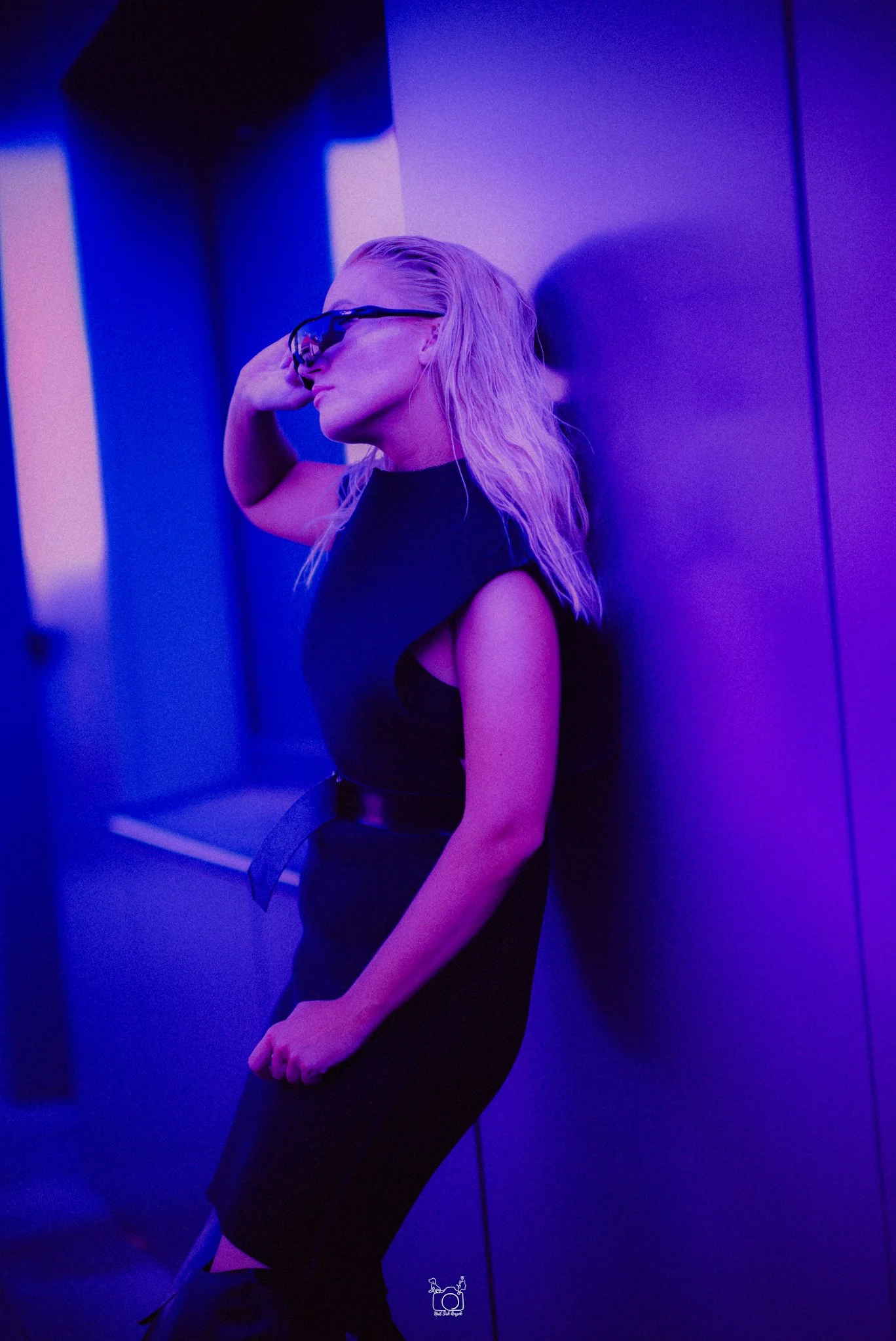 A woman with long blonde hair and sunglasses, wearing a black dress, leaning against a purple wall in a dim, blue-lit environment.