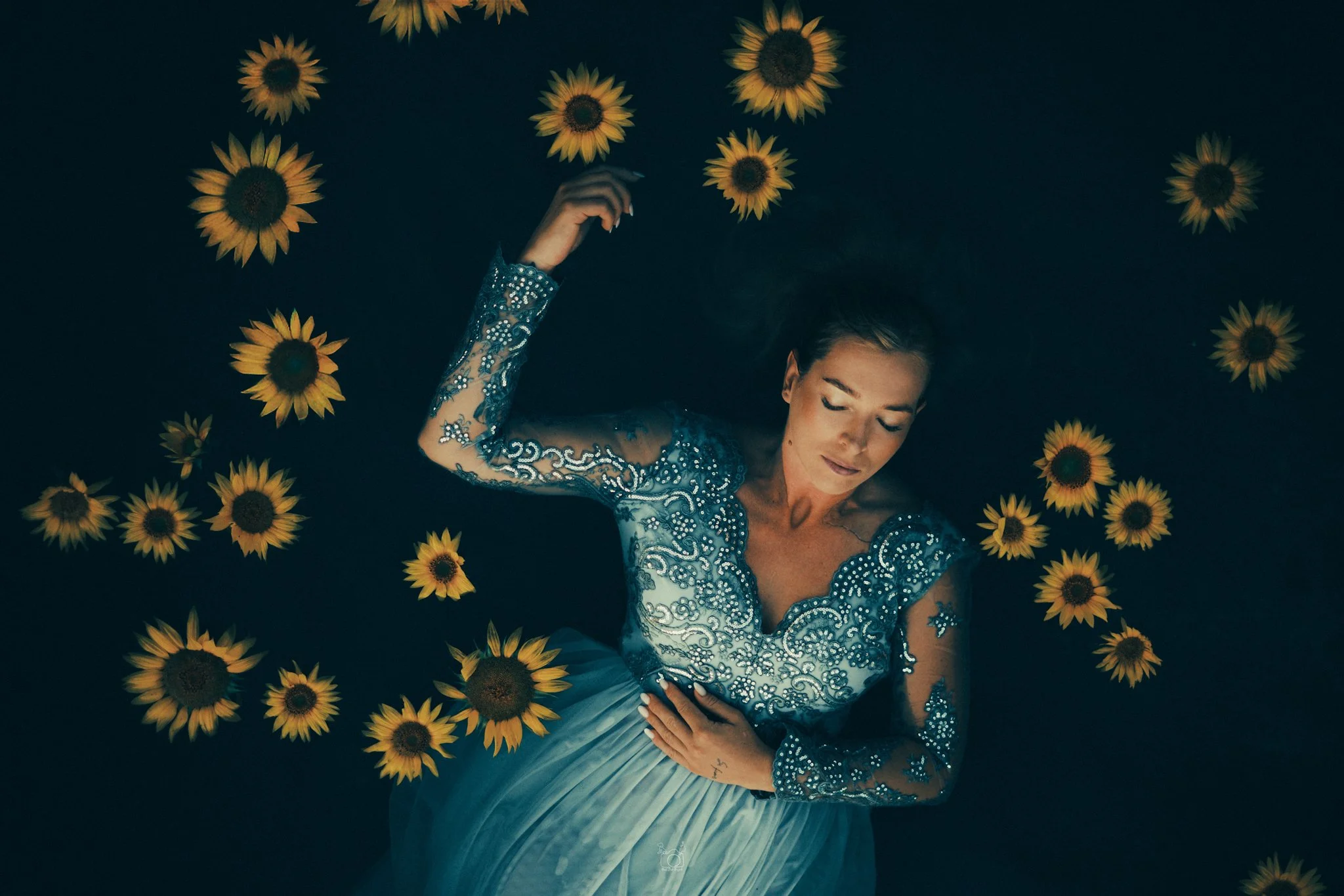 A woman dressed in a blue, beaded gown lying on a black background decorated with yellow sunflowers, with her eyes closed and hand on her chest.