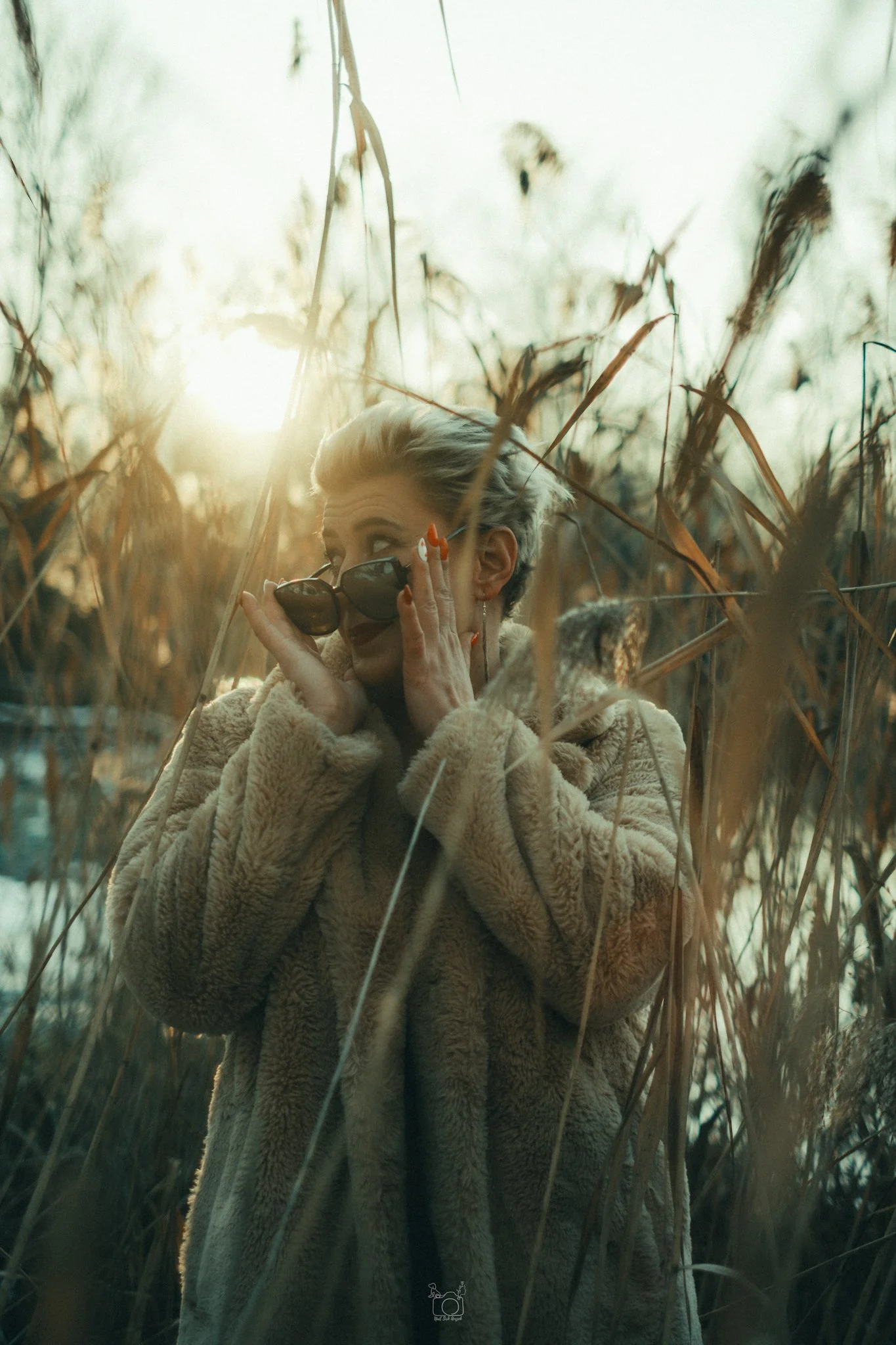 A woman with short, gray hair wearing sunglasses and a beige, fuzzy coat stands amidst tall dry reeds, sunlight shining behind her.