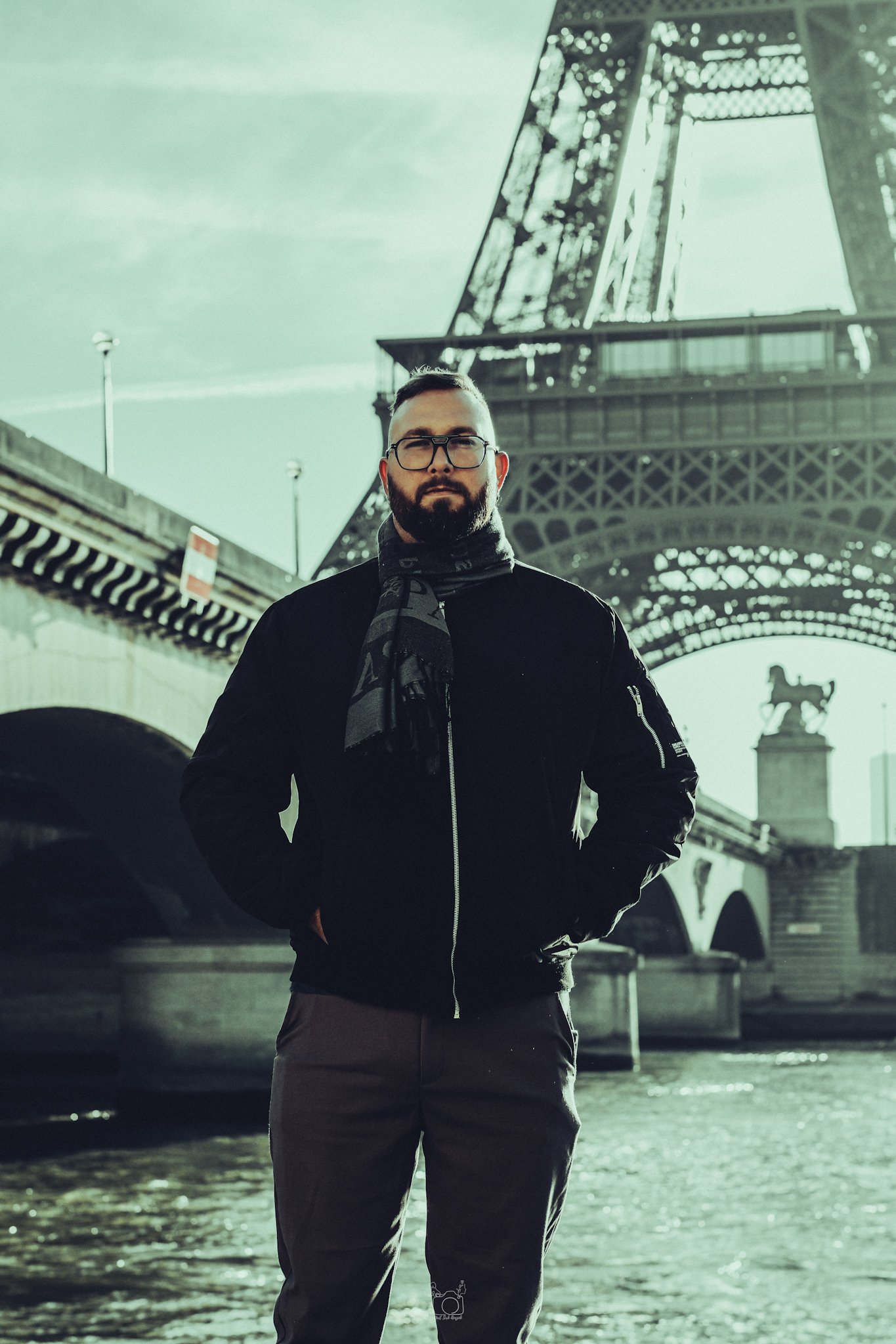 A man with a beard, glasses, and a black jacket stands under the Eiffel Tower during daylight, with water and cars in the background.