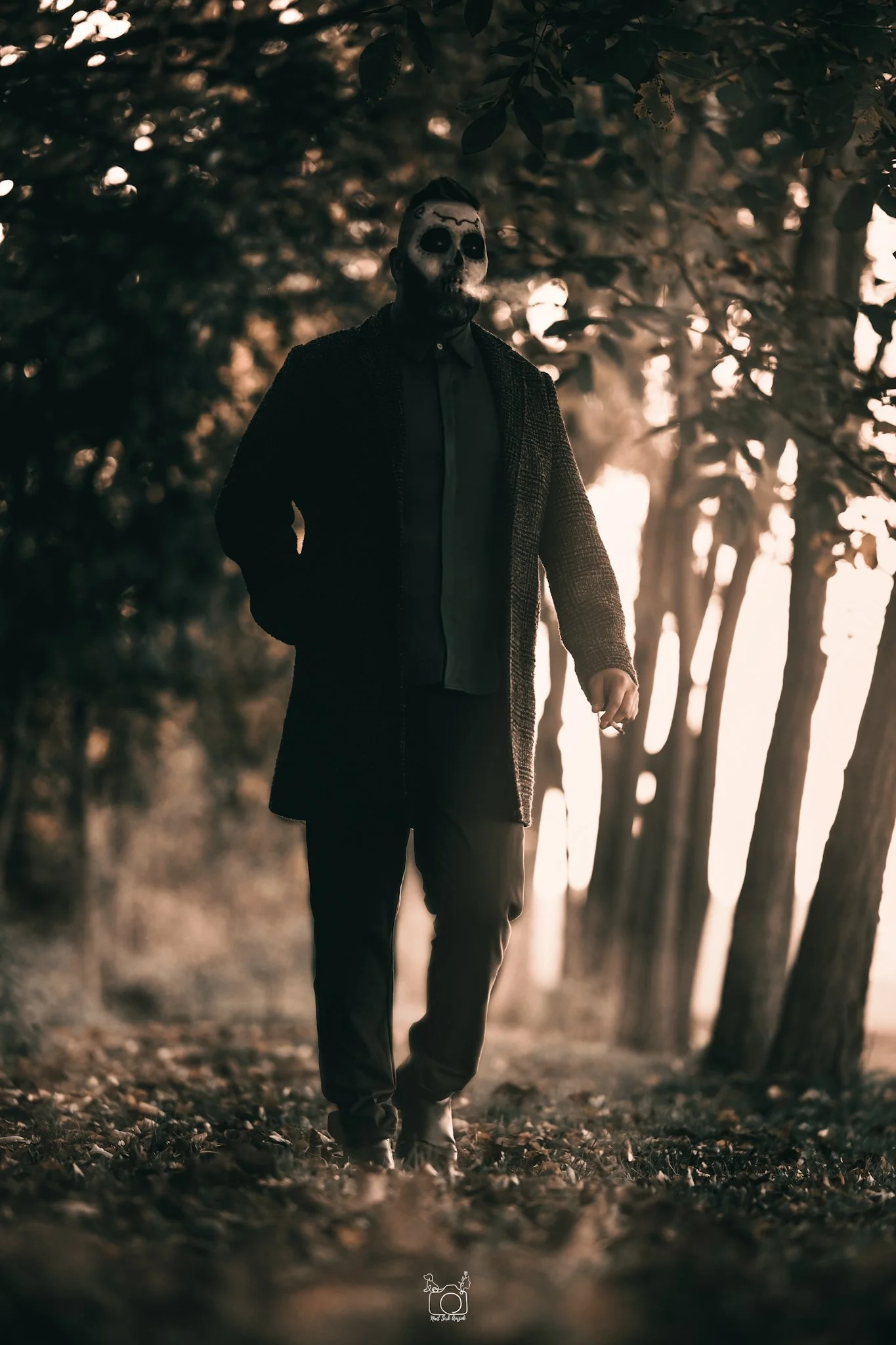A person hiking through a forest at sunset, wearing a mask with dark eye sockets and a beard, dressed in a dark coat and pants. The scene is illuminated by warm, golden sunlight filtering through the trees.