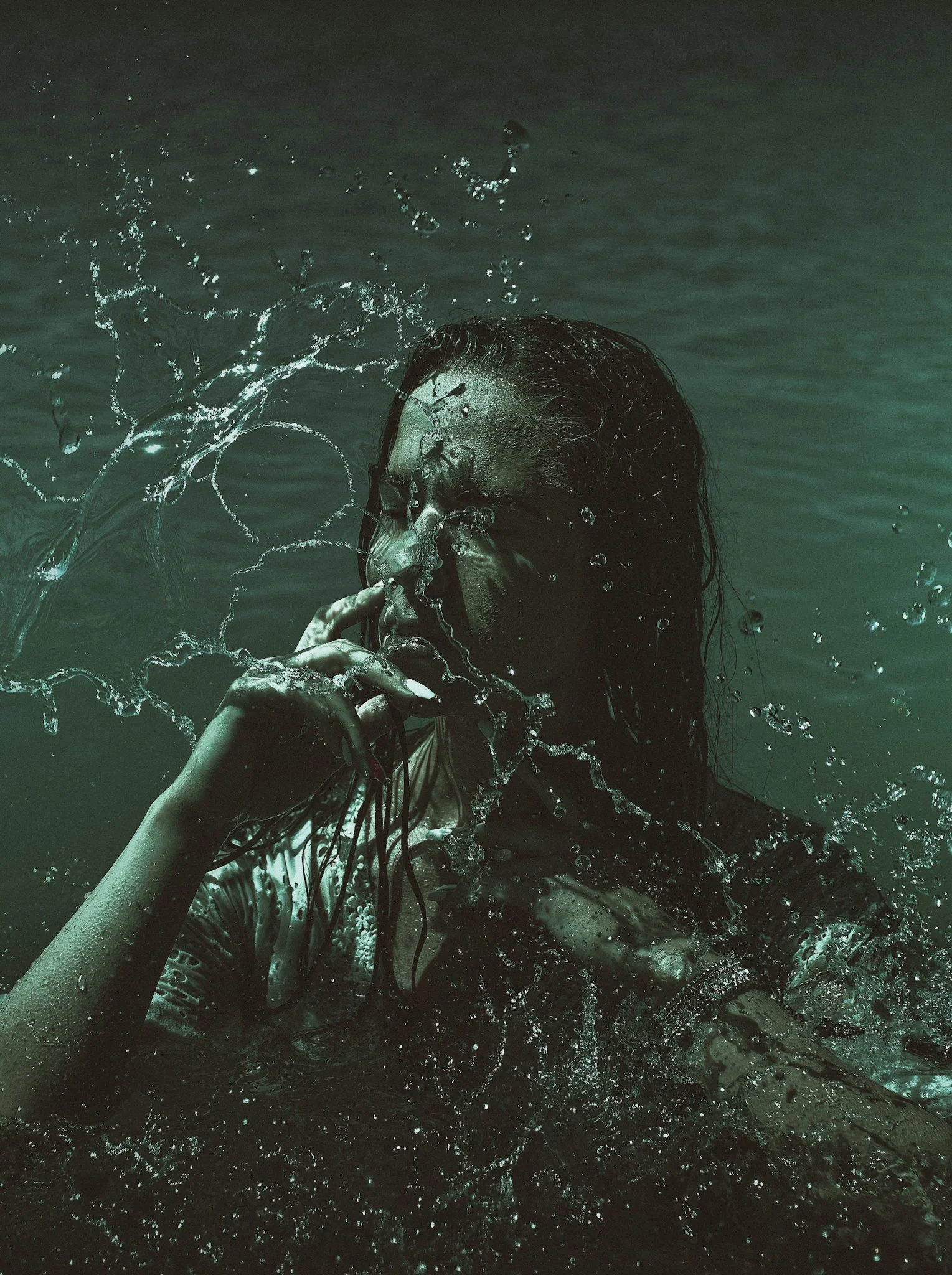 A person with long dark wet hair and sunglasses in water, surrounded by splashing water droplets.