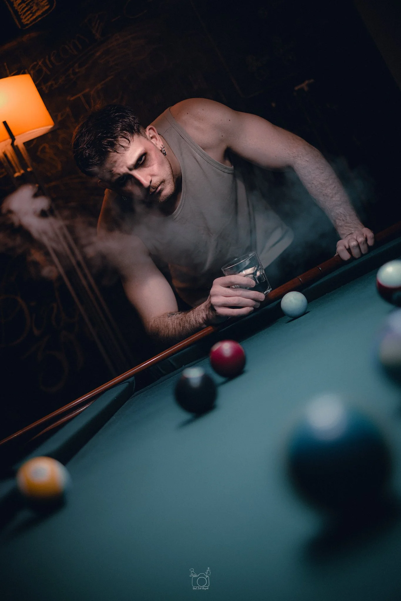 A man with dark hair, wearing a sleeveless shirt, leans over a pool table, holding a glass of drink, with pool balls on the table and a lamp with a beige shade in the background.