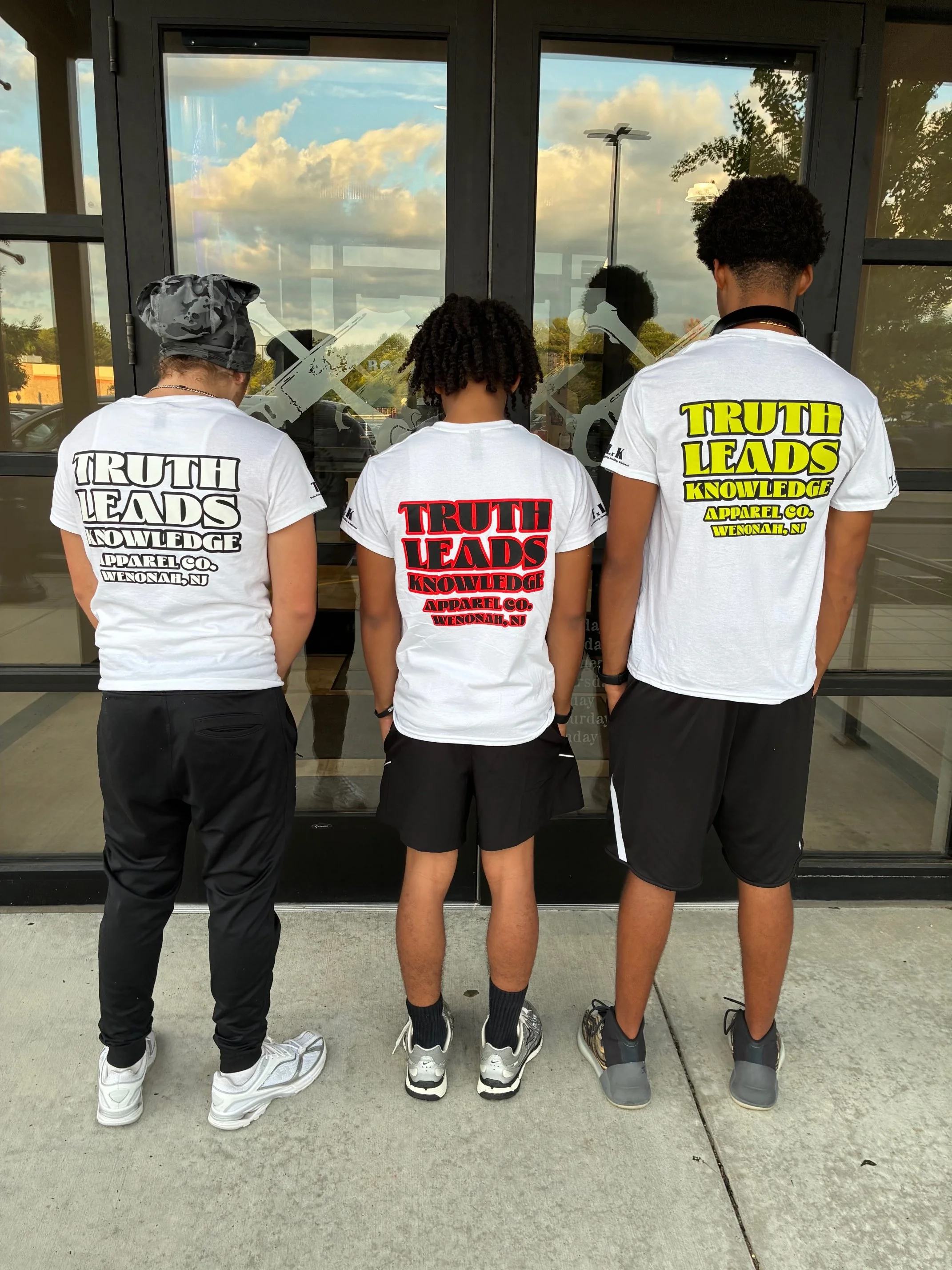 Three people standing with their backs to the camera, wearing matching white T-shirts with bold text on the back, standing outside in front of glass doors that reflect trees and a cloudy sky.