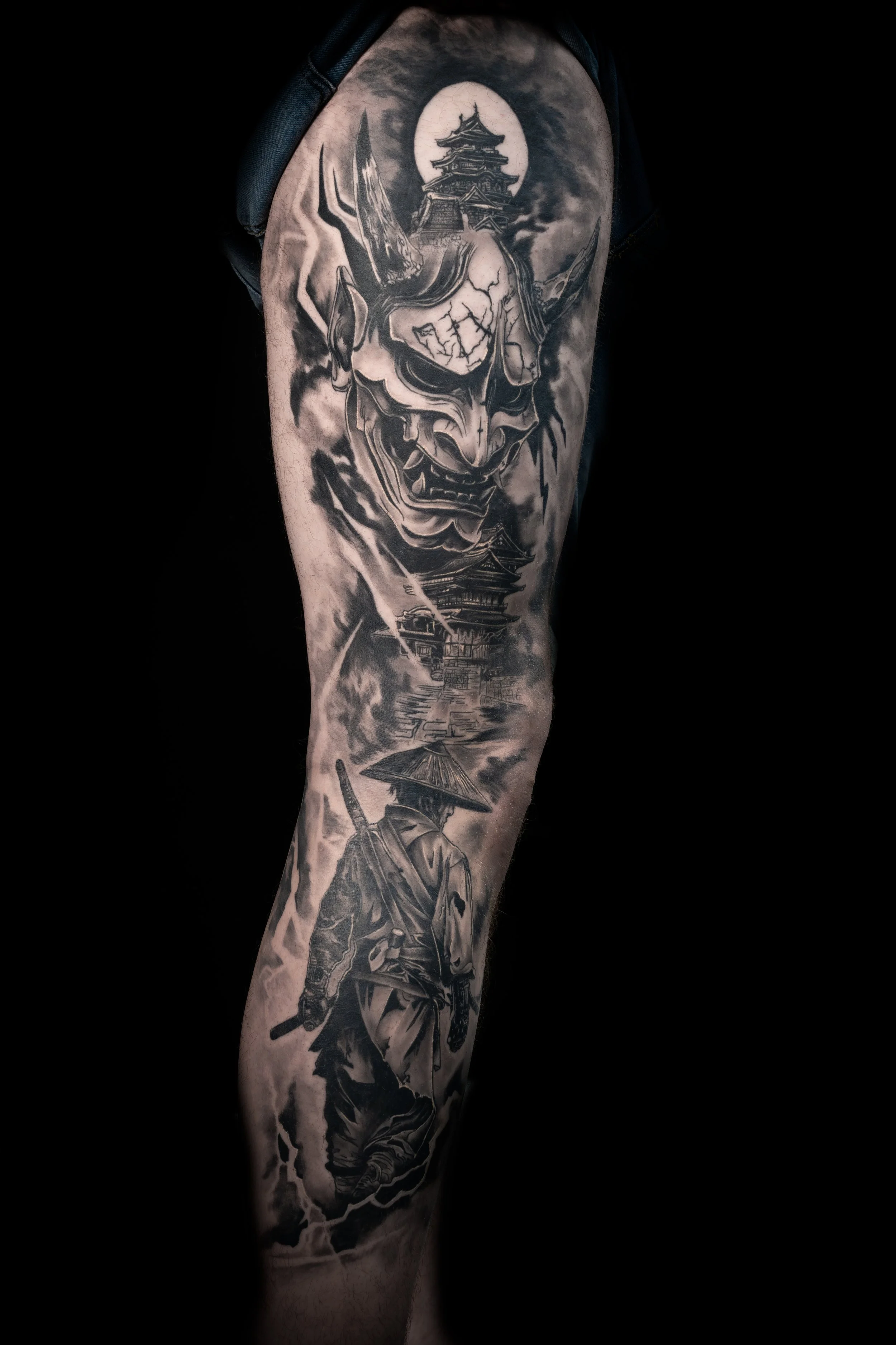 Tattoo of a fierce demon face with sharp features, a temple and mountainscape in the background, on a person's upper arm.