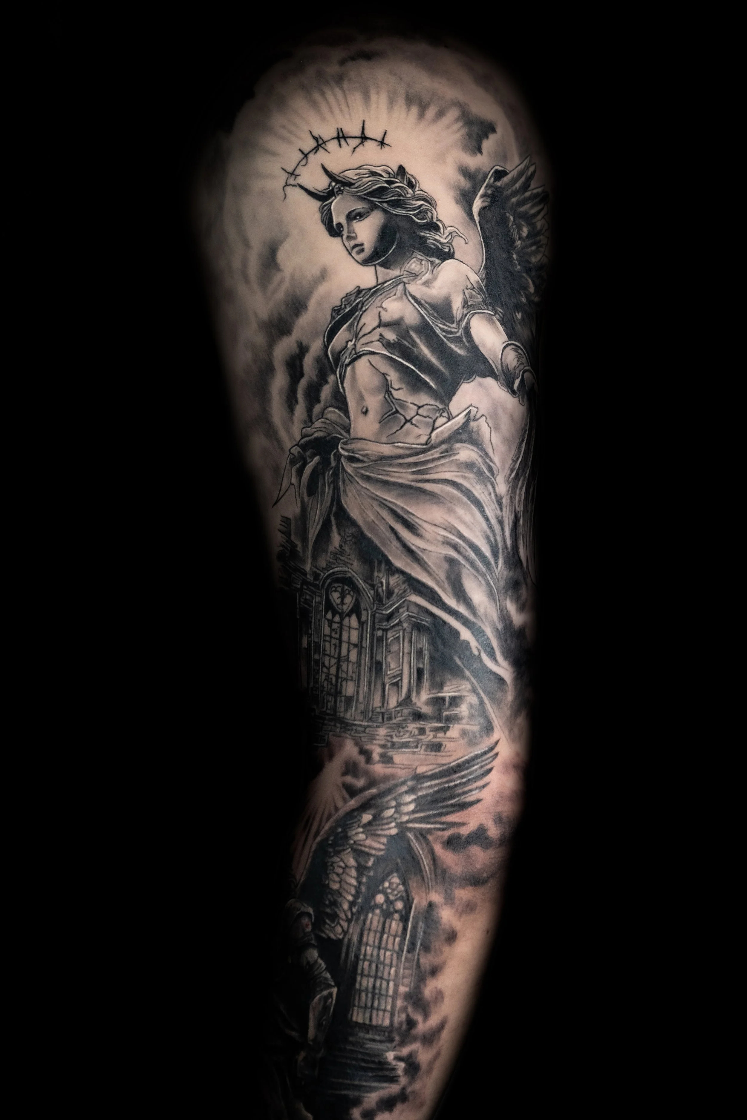 Black and gray detailed tattoo of a woman with wings, a horn, and a halo, with a cathedral and waves below, on a person's arm.