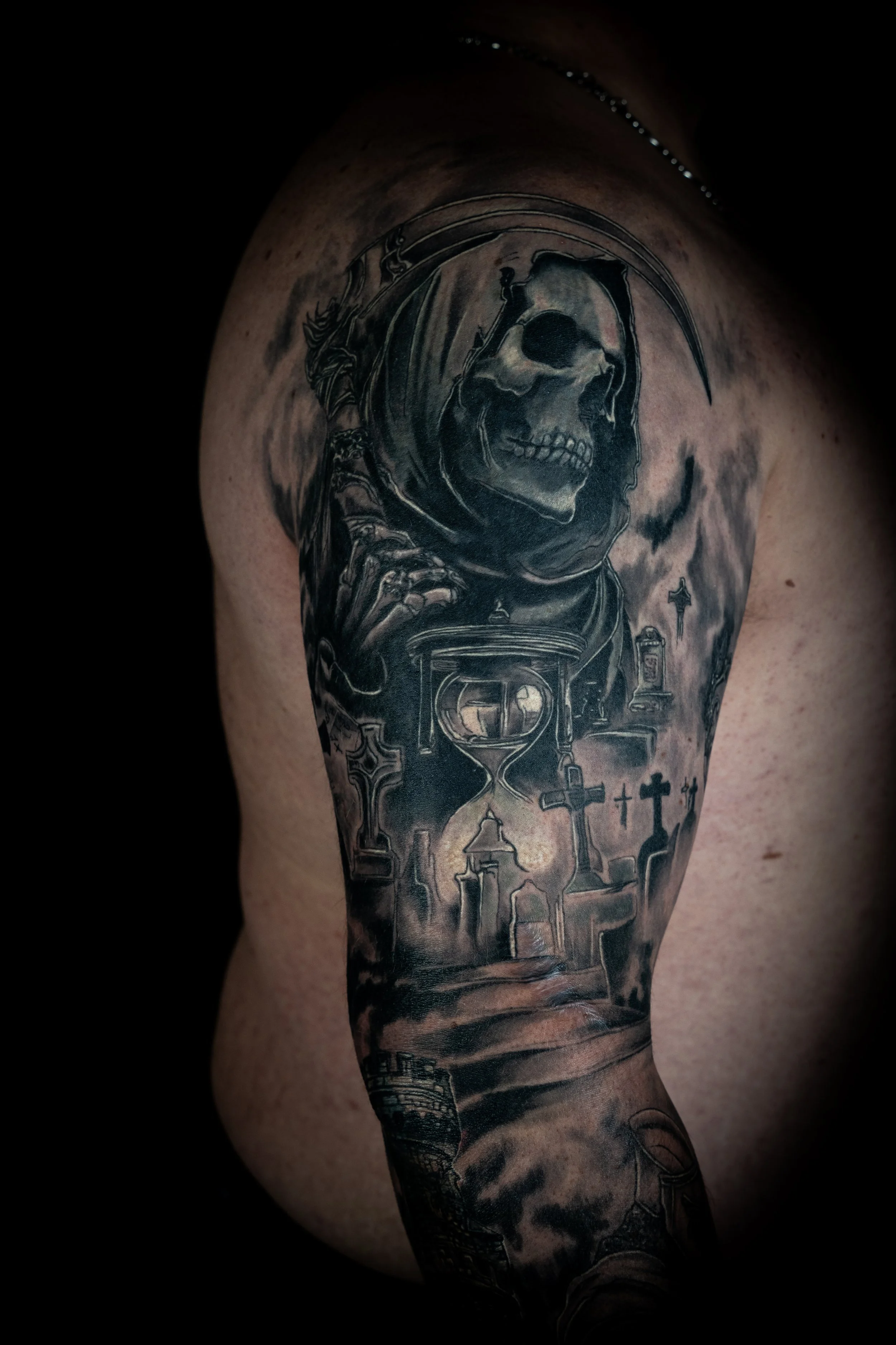 Black and gray tattoo of a skull with a scythe inside a circular frame, with various other elements below, on a person's upper arm.