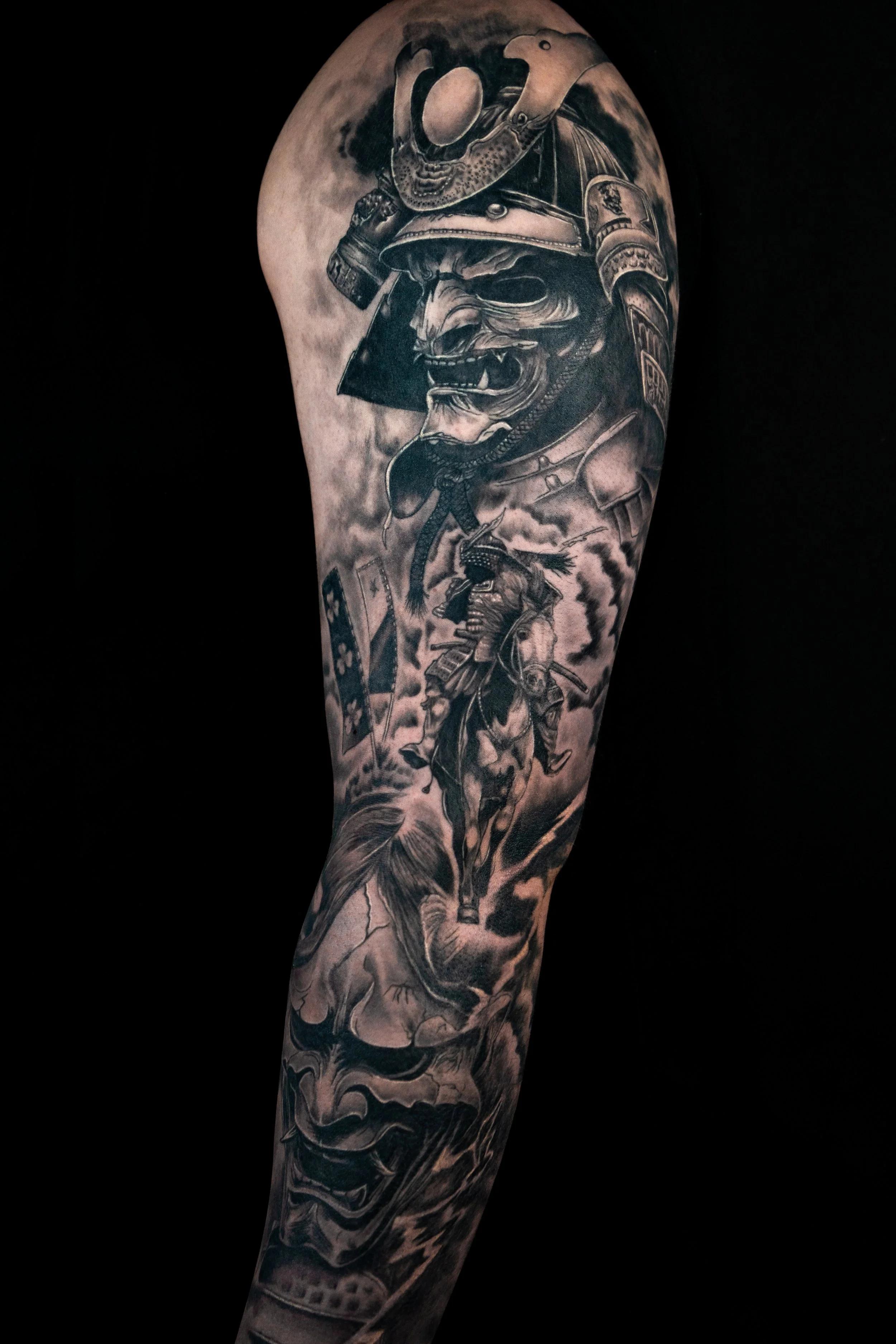 Black and gray tattoo sleeve featuring a fierce, snarling dog wearing a samurai helmet, surrounded by clouds and smoke effects.