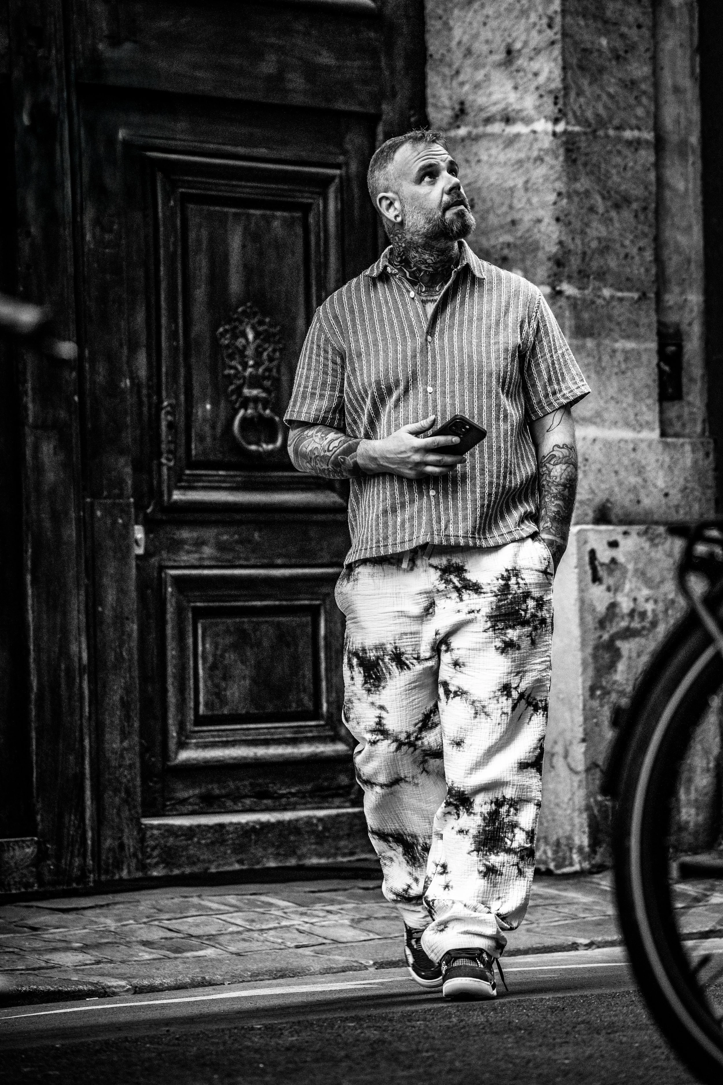 Un homme avec des tatouages porte un smartphone dans la main et marche à côté d'une rue pavée devant une porte en bois.