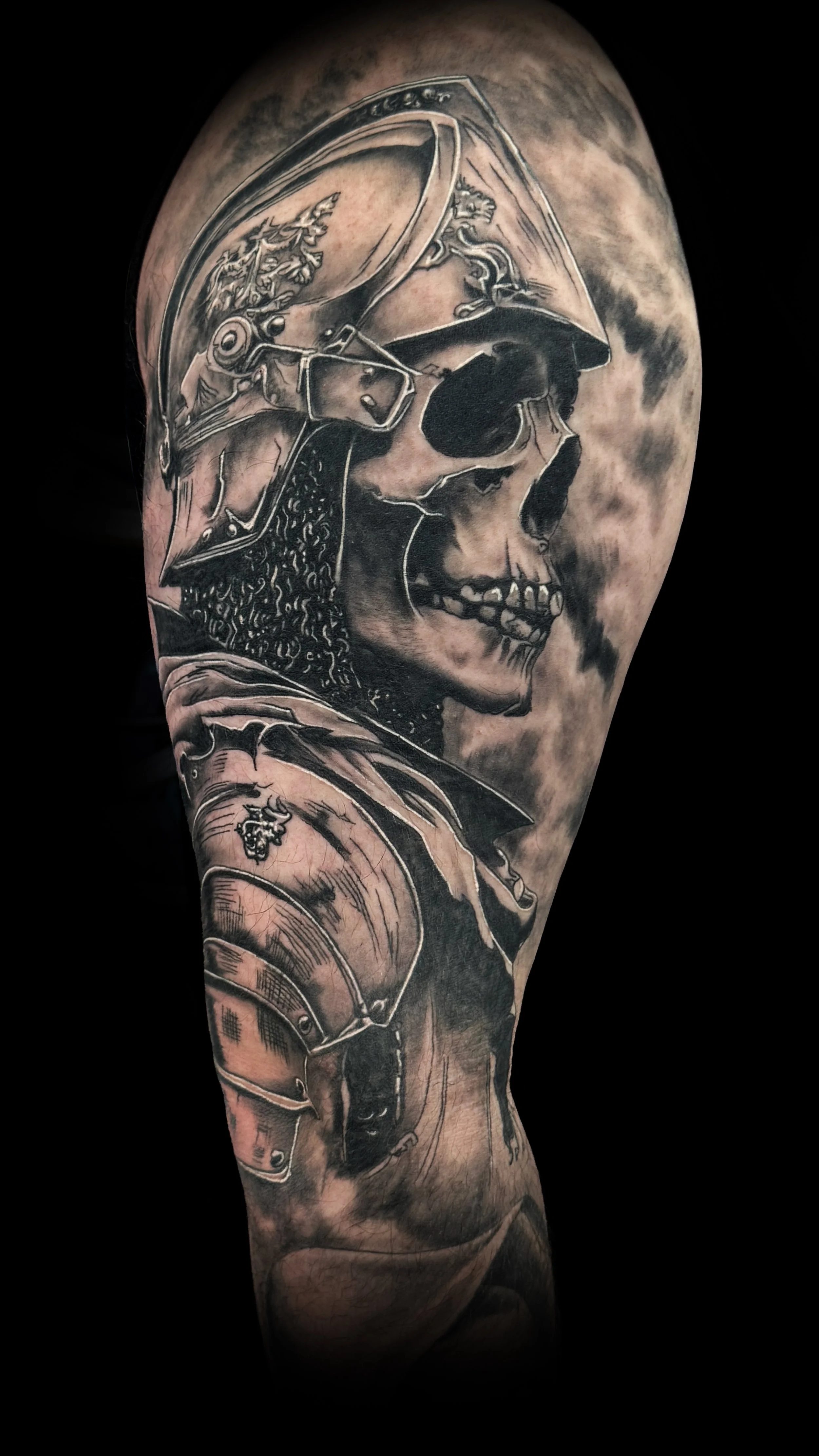Black and grey tattoo of a skull wearing a military helmet with a chainmail beard on a person's arm.