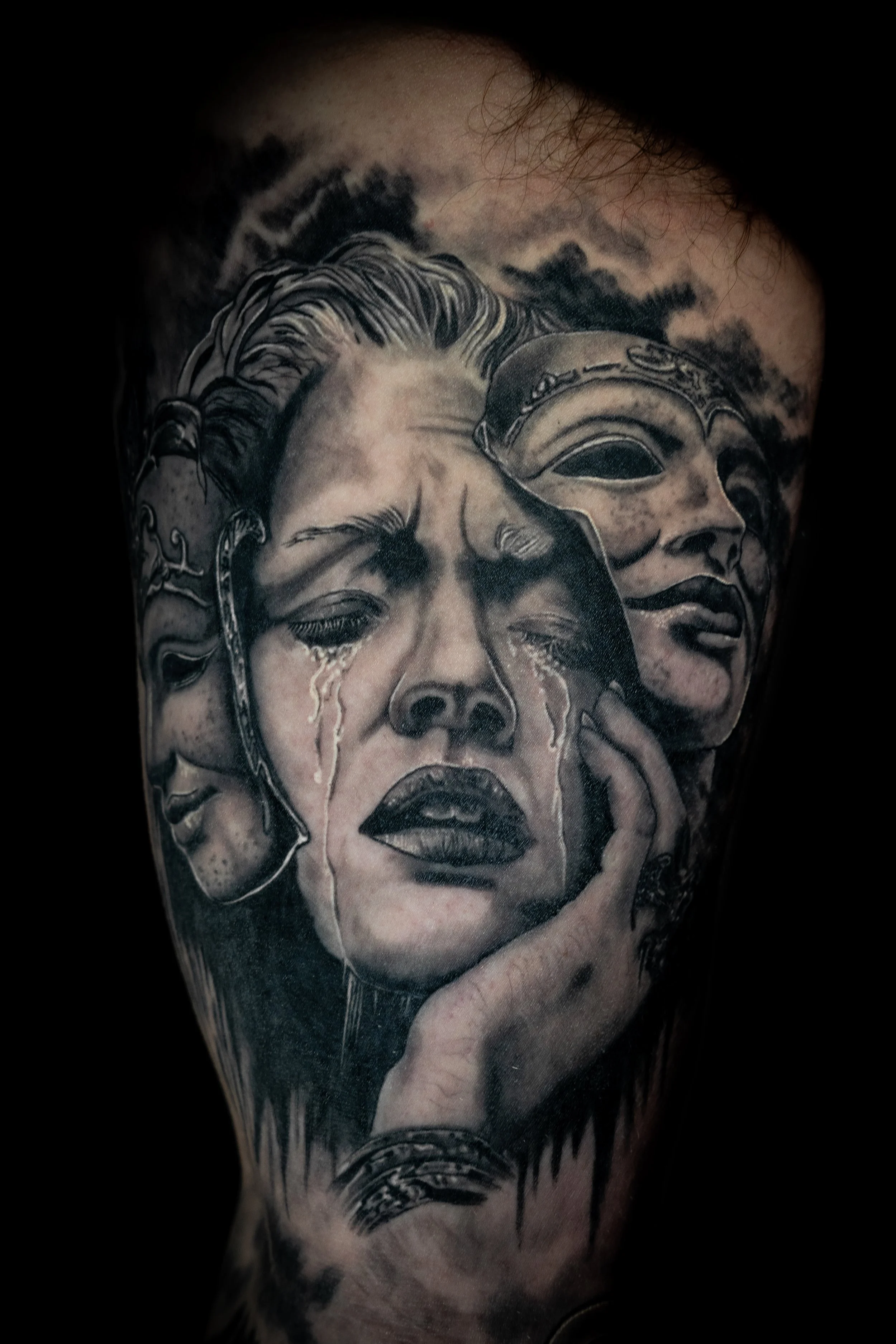 A black and gray tattoo depicting multiple female faces with different expressions, some crying, with detailed shading and intricate designs around them.