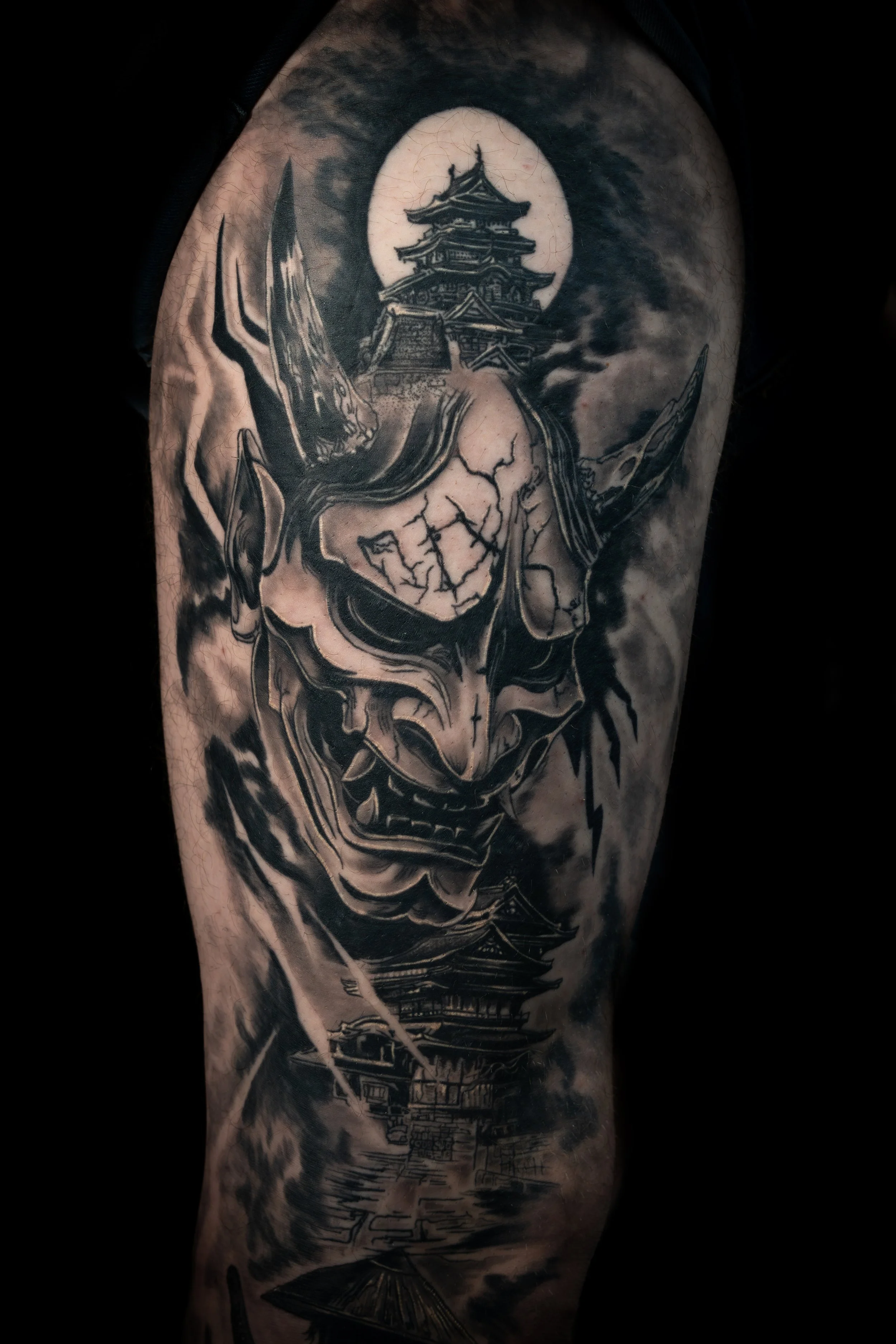 Black and grey tattoo of a traditional Japanese pagoda on a person's upper arm, with a full moon in the background and abstract swirling lines.