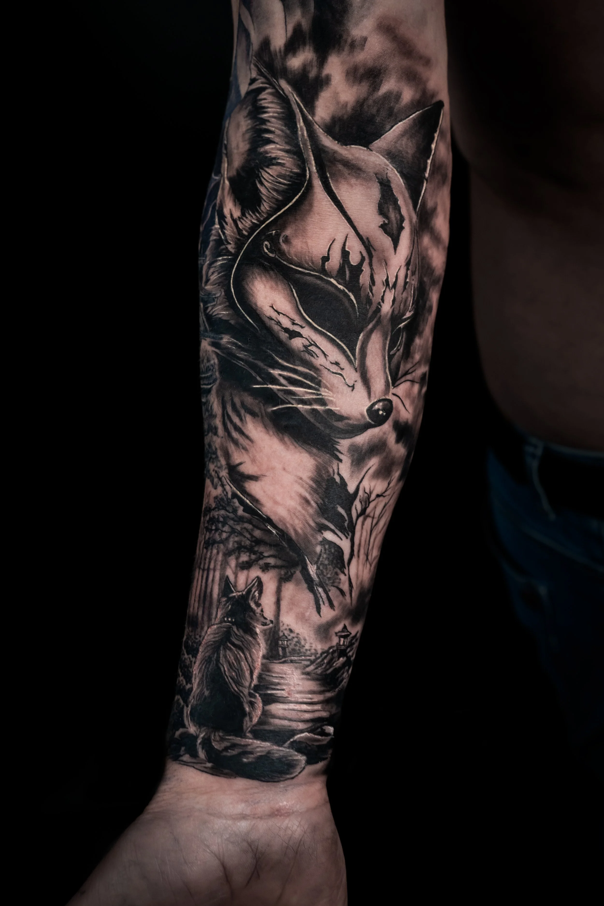 Tattoo sleeve featuring a wolf howling by a lake, with trees and buildings in the background, in black and gray ink.