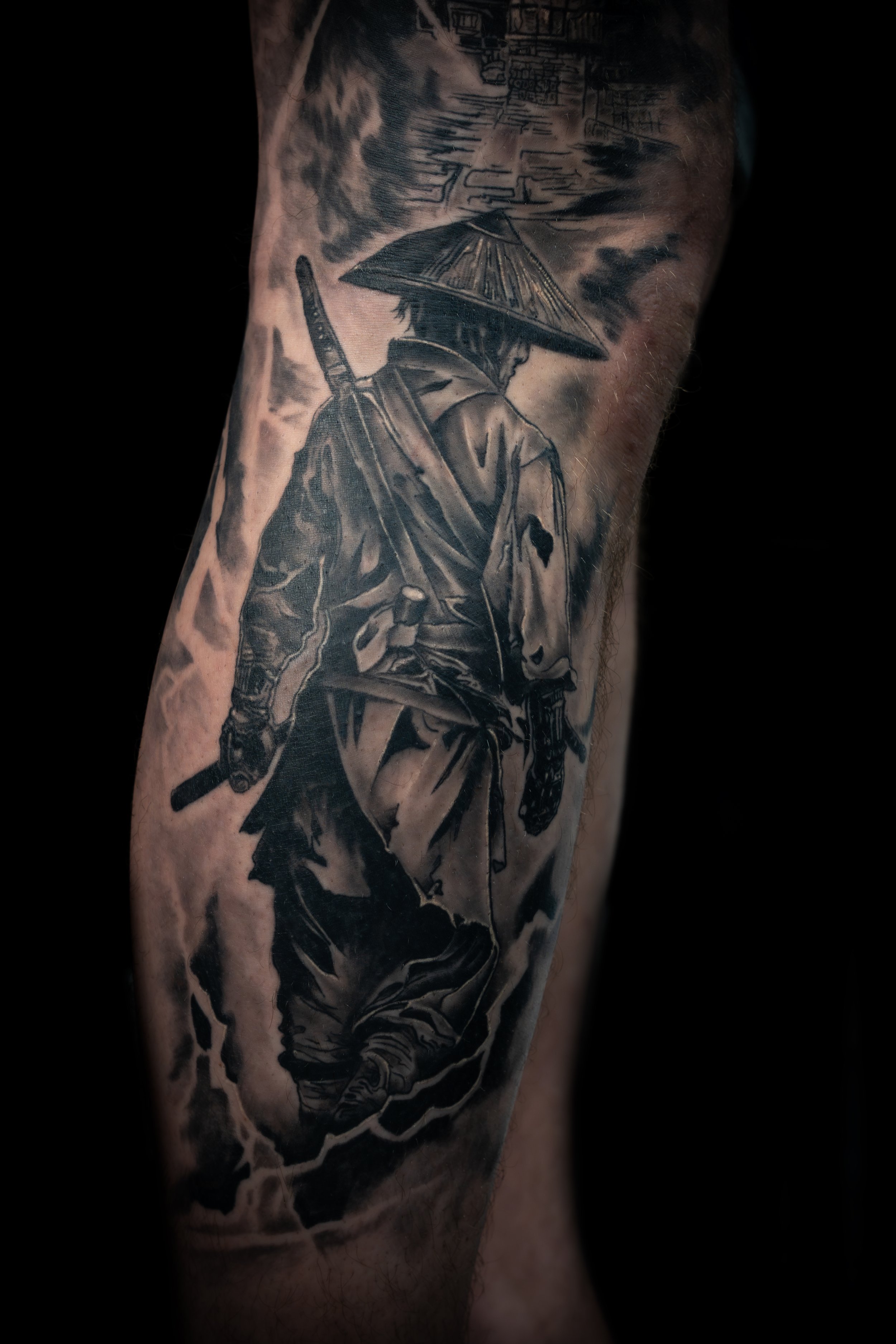 Black and gray tattoo of a samurai warrior on a person's arm, wearing a traditional hat and carrying a sword, with a background of clouds and landscapes.