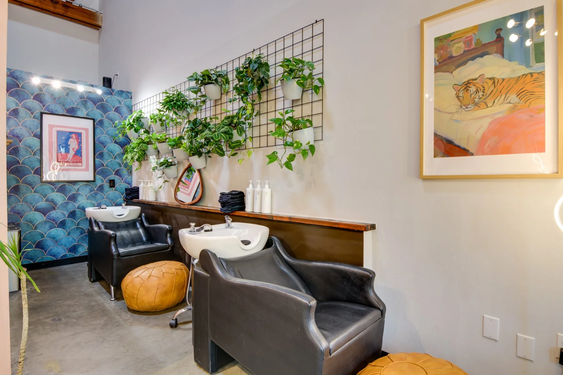Hair salon with two shampoo sinks, black salon chairs, a pouf, and decorative wall art, including framed artwork and potted plants on a grid wall.