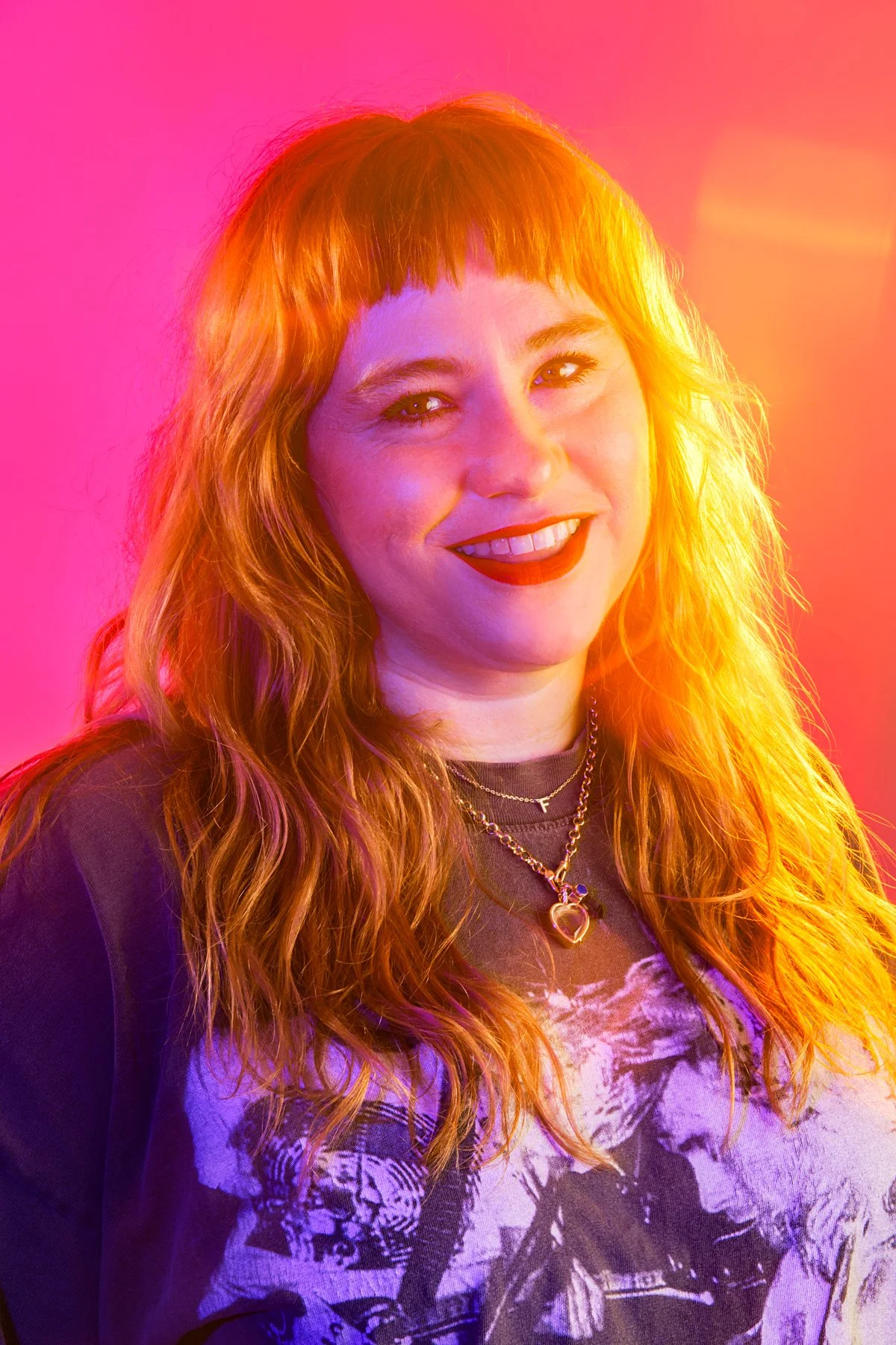 A woman with red hair smiling, wearing a black t-shirt with a graphic print, gold necklaces, and a dark jacket, illuminated by colorful pink, purple, and yellow lighting.