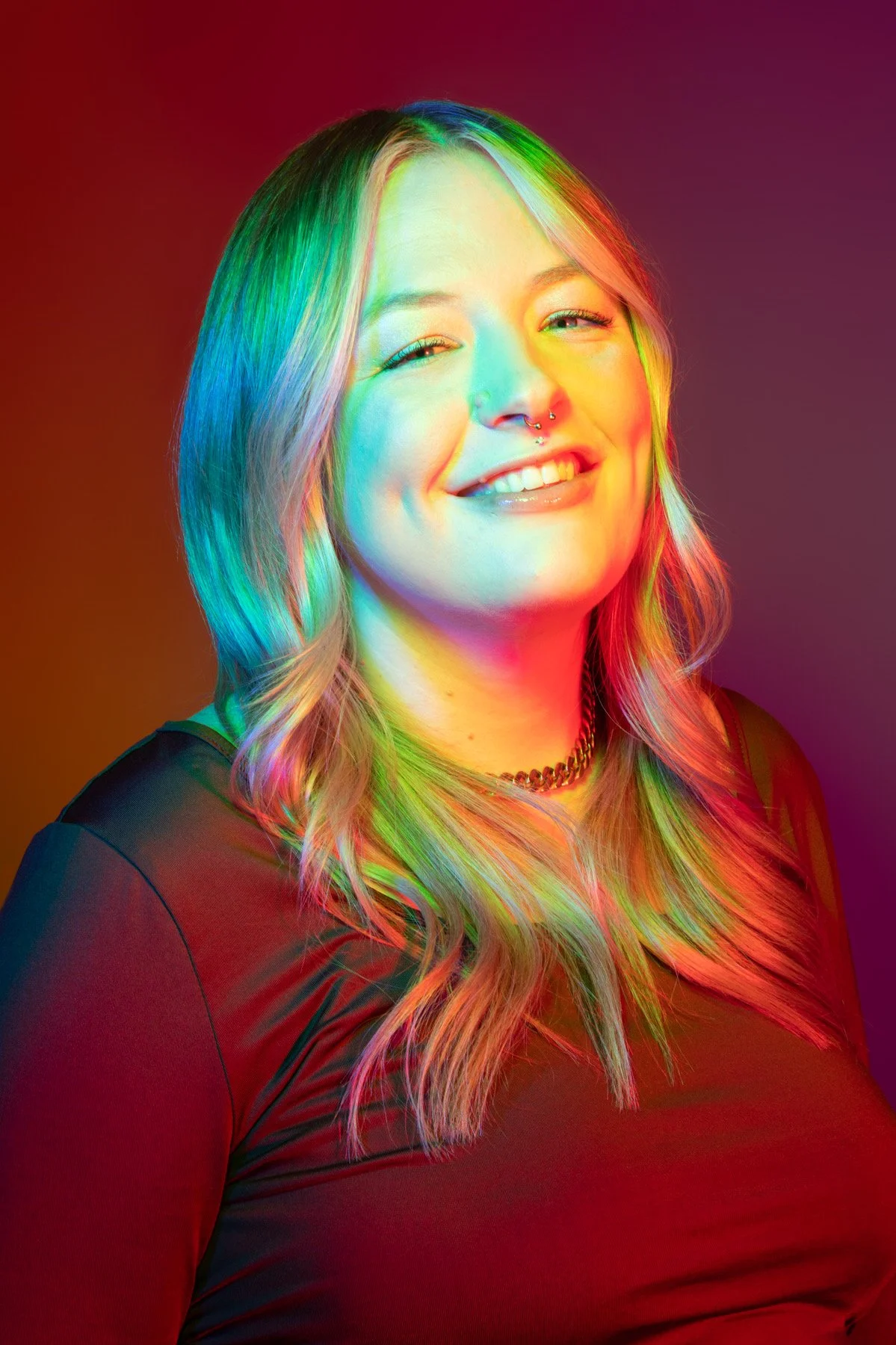 A woman with long, wavy hair and a septum piercing, smiling with closed eyes against a colorful rainbow gradient background.