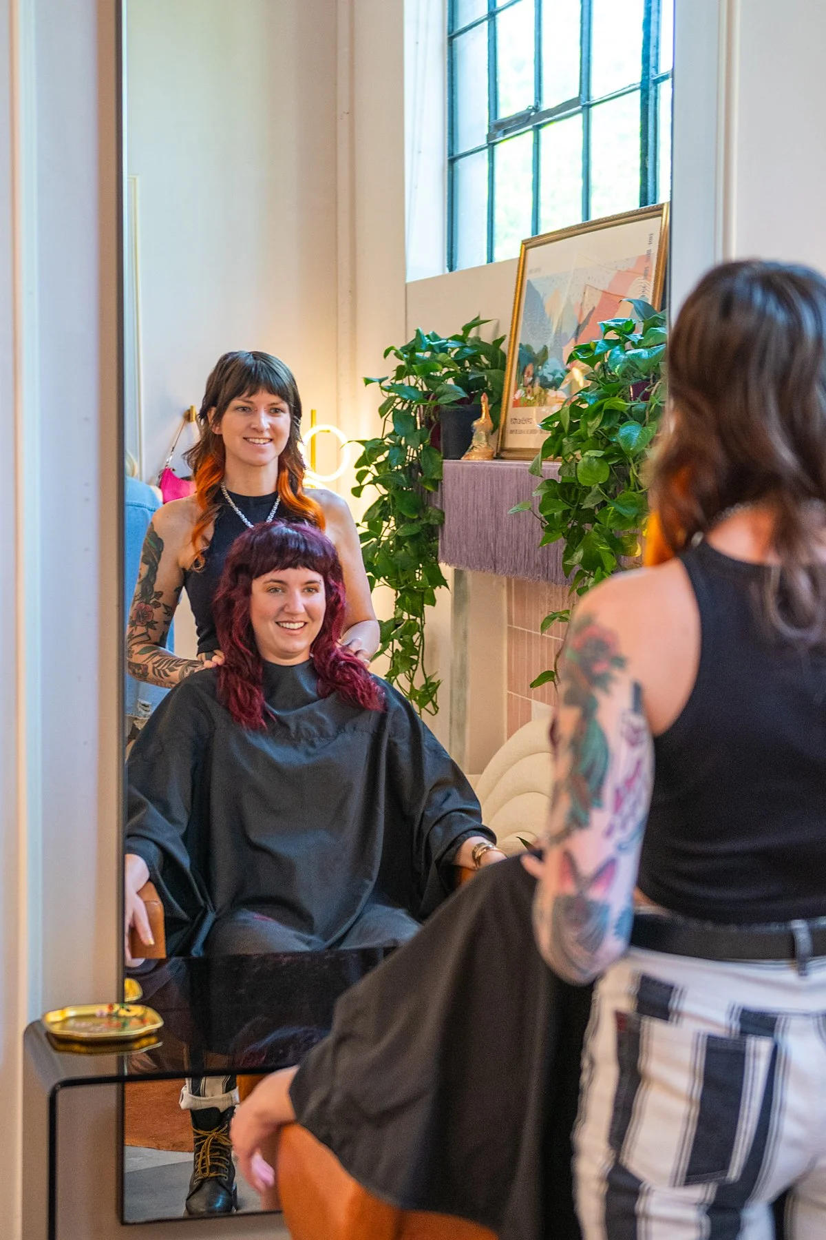 Two women with tattoos getting ready for a haircut in a salon, reflected in a mirror. One woman is seated with a black cape, and the other woman, with tattoos on her arms and wearing a black top, is standing.