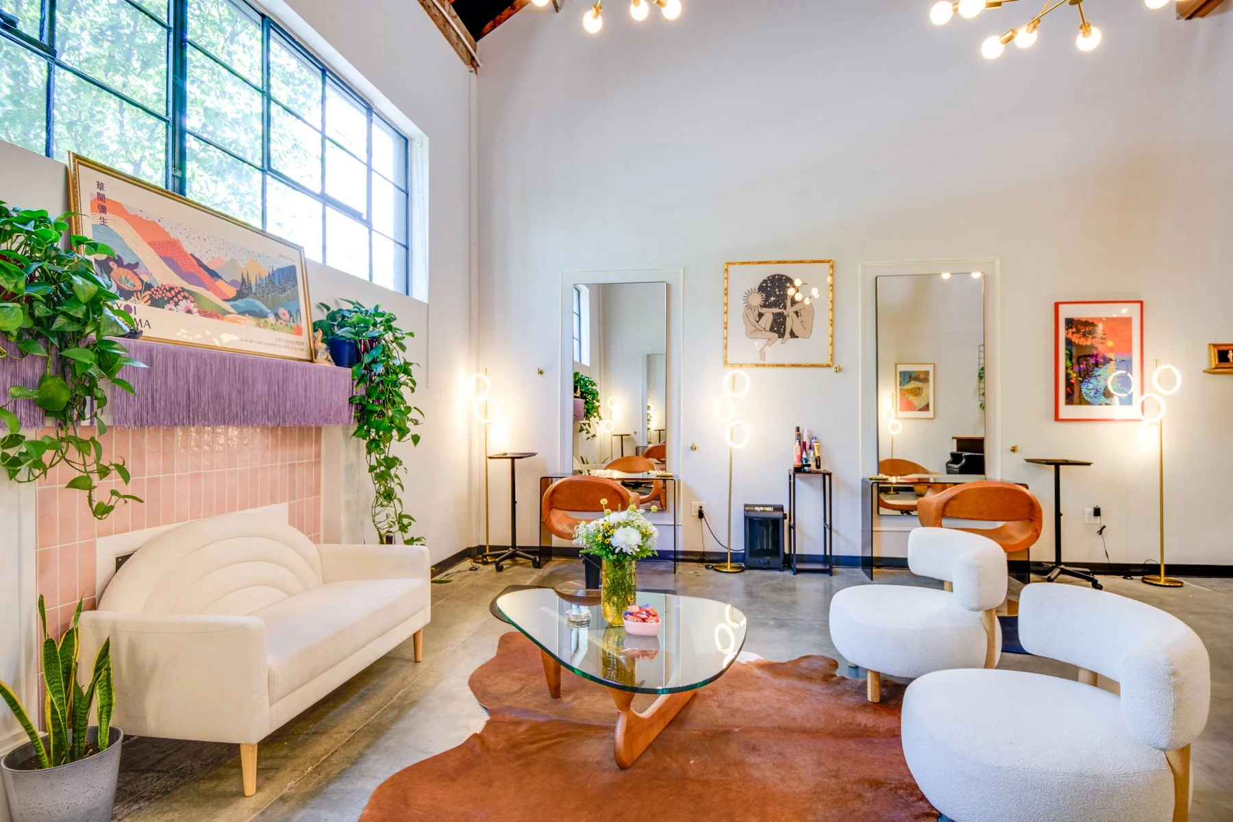 A cozy interior with modern decor, including a white sofa, patterned rug, glass coffee table with a flower vase, and multiple chairs. Bright artwork and plants decorate the walls, and there are standing lamps with circular light fixtures. Large window with a grid pattern allows natural light into the space.