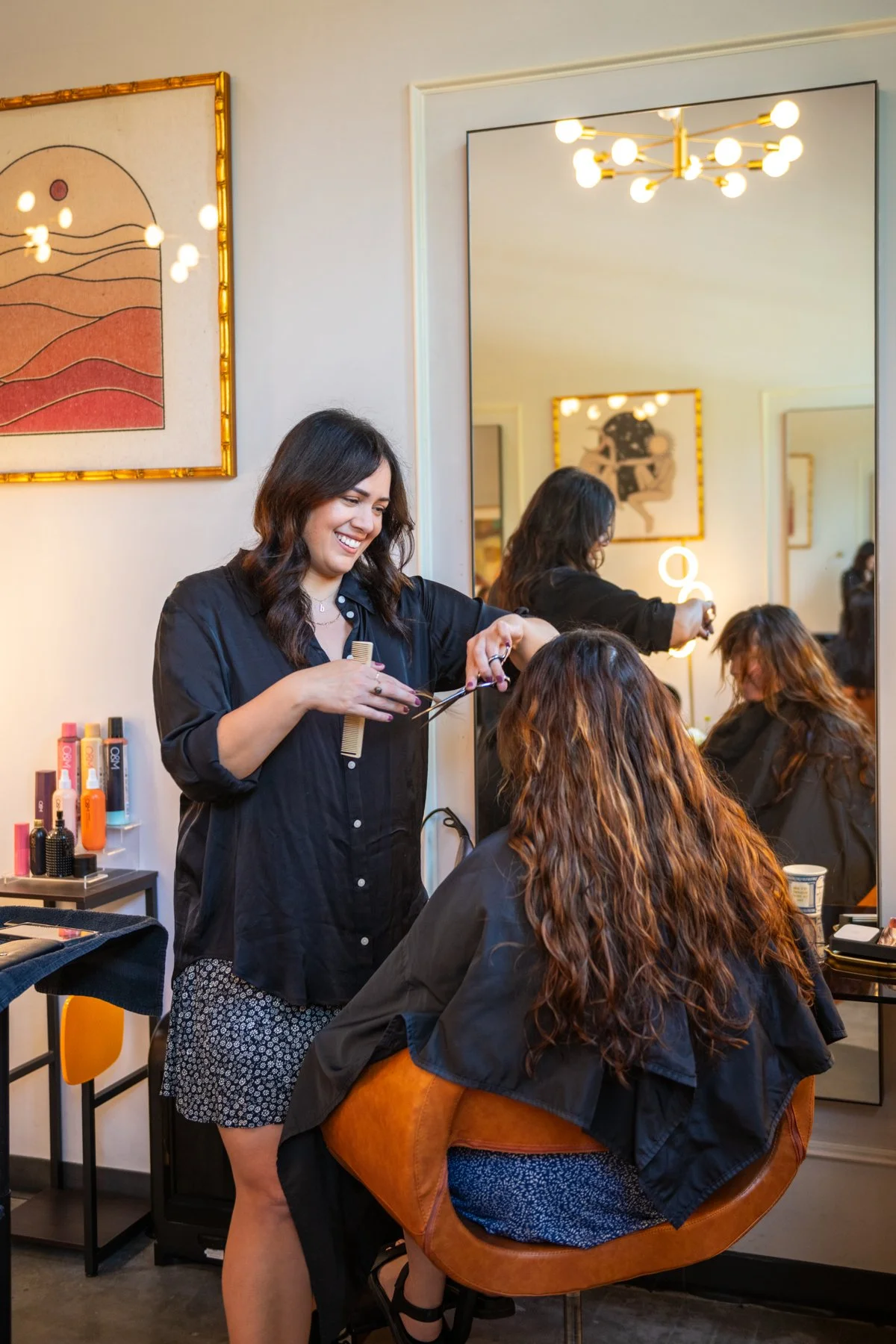 A hairstylist with dark, wavy hair and a black button-up shirt is styling a woman's long, curly hair in a salon. The woman is seated in a brown, upholstered chair, wearing a black cape. The salon has a large mirror, colorful artworks, and hair care products on nearby shelves. There is warm indoor lighting and a modern chandelier.