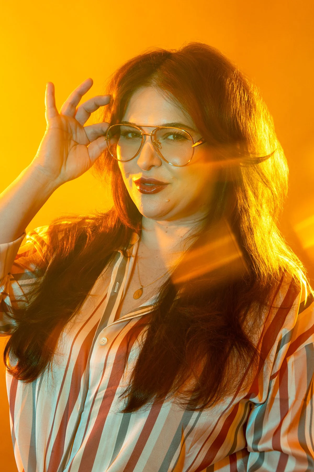 A woman with long dark hair wearing glasses, gold jewelry, and a striped shirt, posing with a yellow-orange background illuminated by strong lighting.