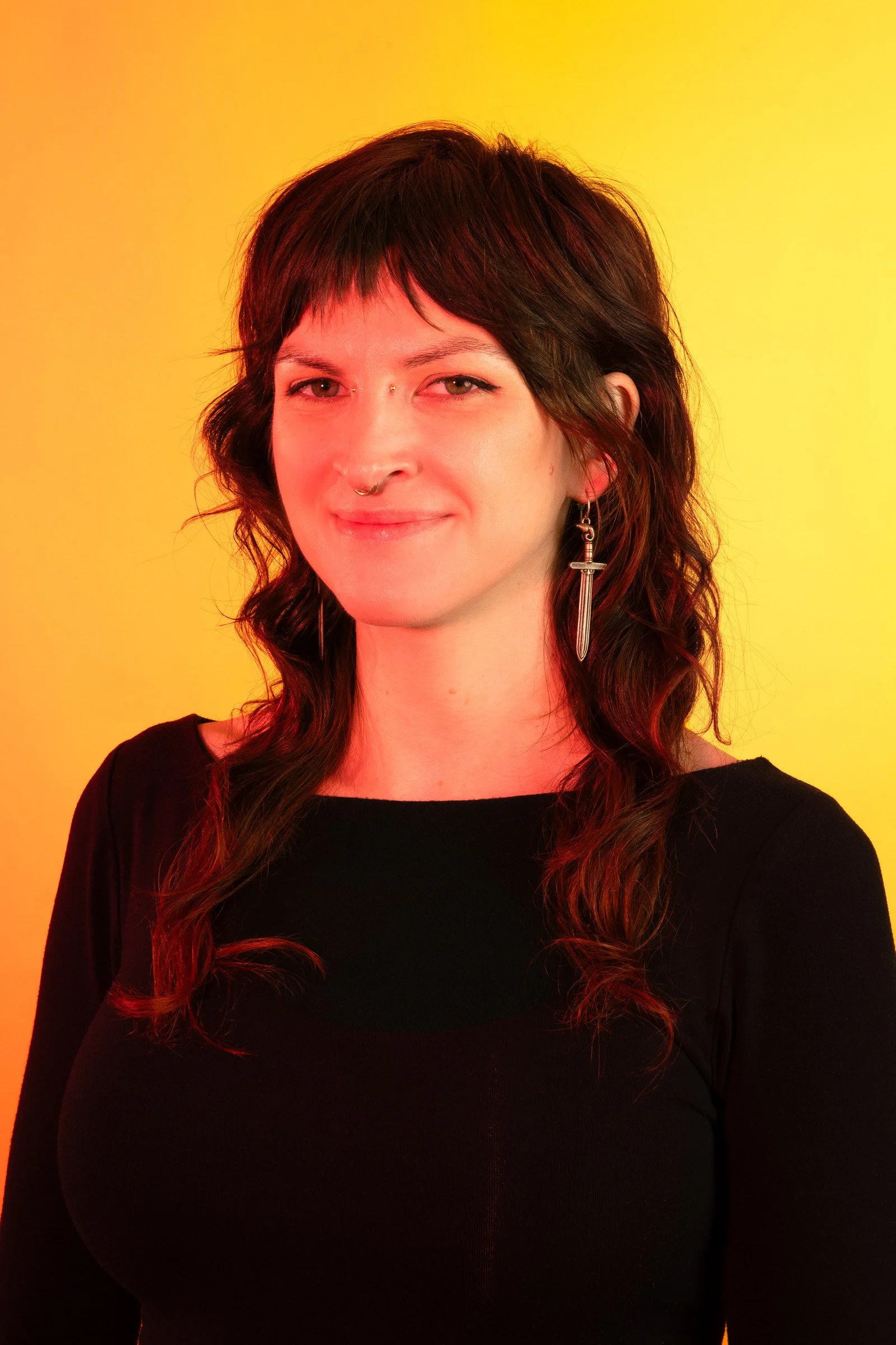 A woman with dark brown, wavy hair and light skin wearing a black top and long earring, standing against a yellow and orange gradient background.