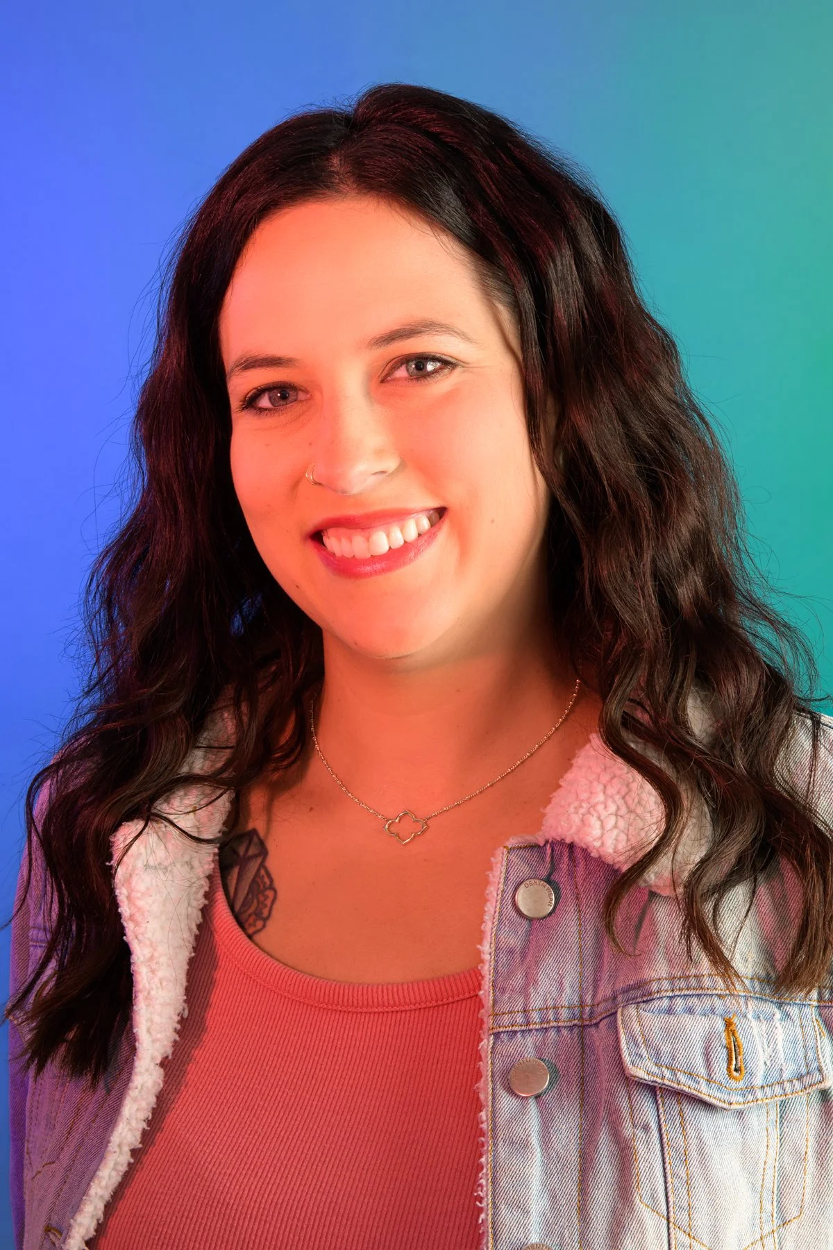 A young woman with dark wavy hair, wearing a denim jacket with a sherpa lining, a coral top, and a gold necklace, smiling against a multicolored background.