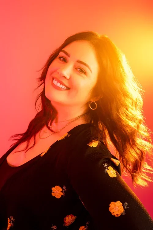 Smiling woman with long, wavy hair, wearing earrings and a black top with floral patterns, illuminated by warm sunlight.