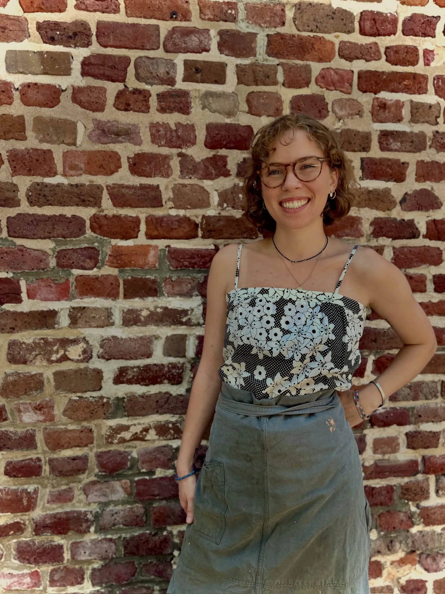 A woman with curly hair and glasses smiling while standing in front of a brick wall, wearing a floral-patterned sleeveless top and a skirt.