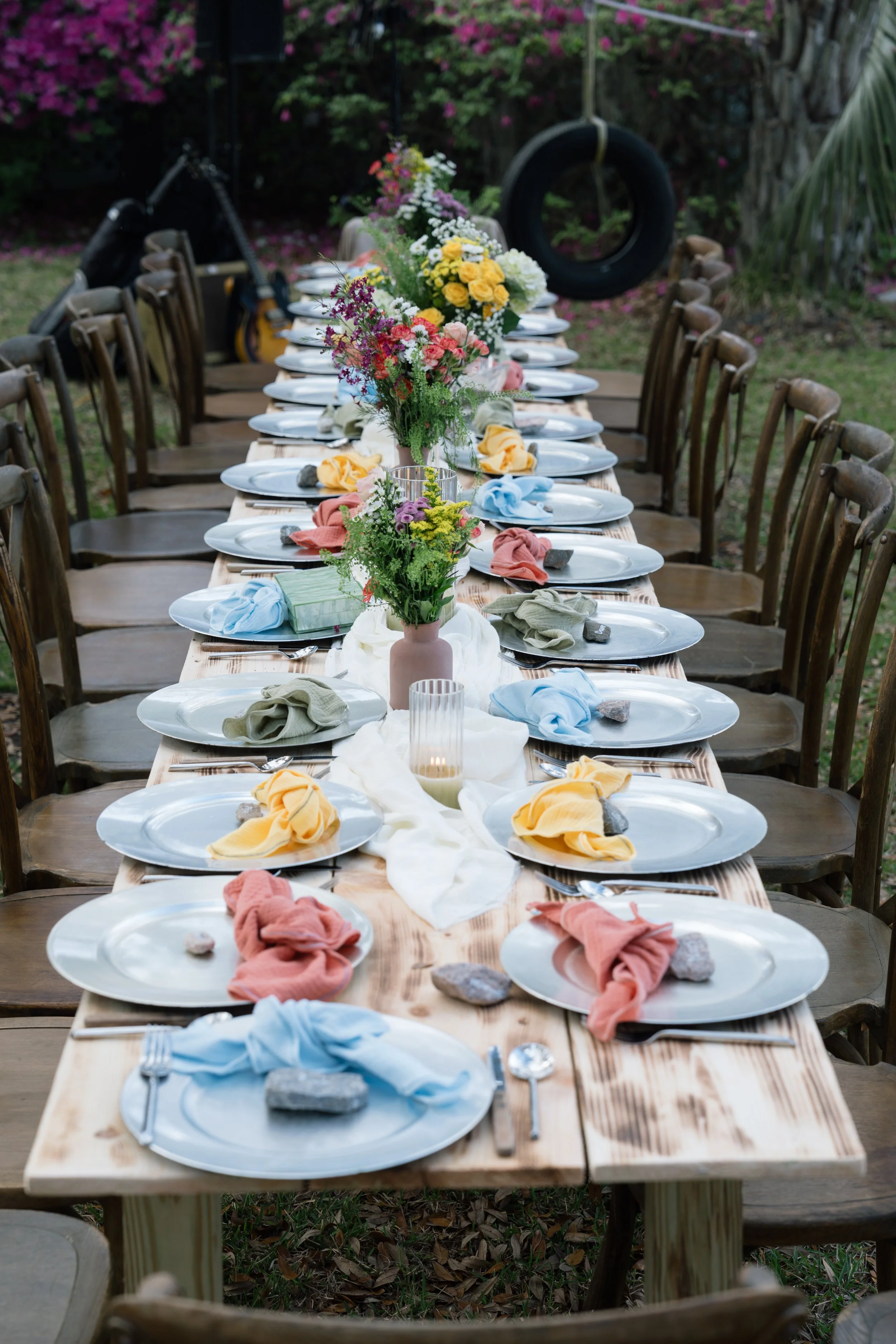 Long wooden outdoor dining table set for a gathering, decorated with colorful flowers, cloth napkins, and stones, surrounded by wooden chairs, with a swing hanging from a tree in the background.