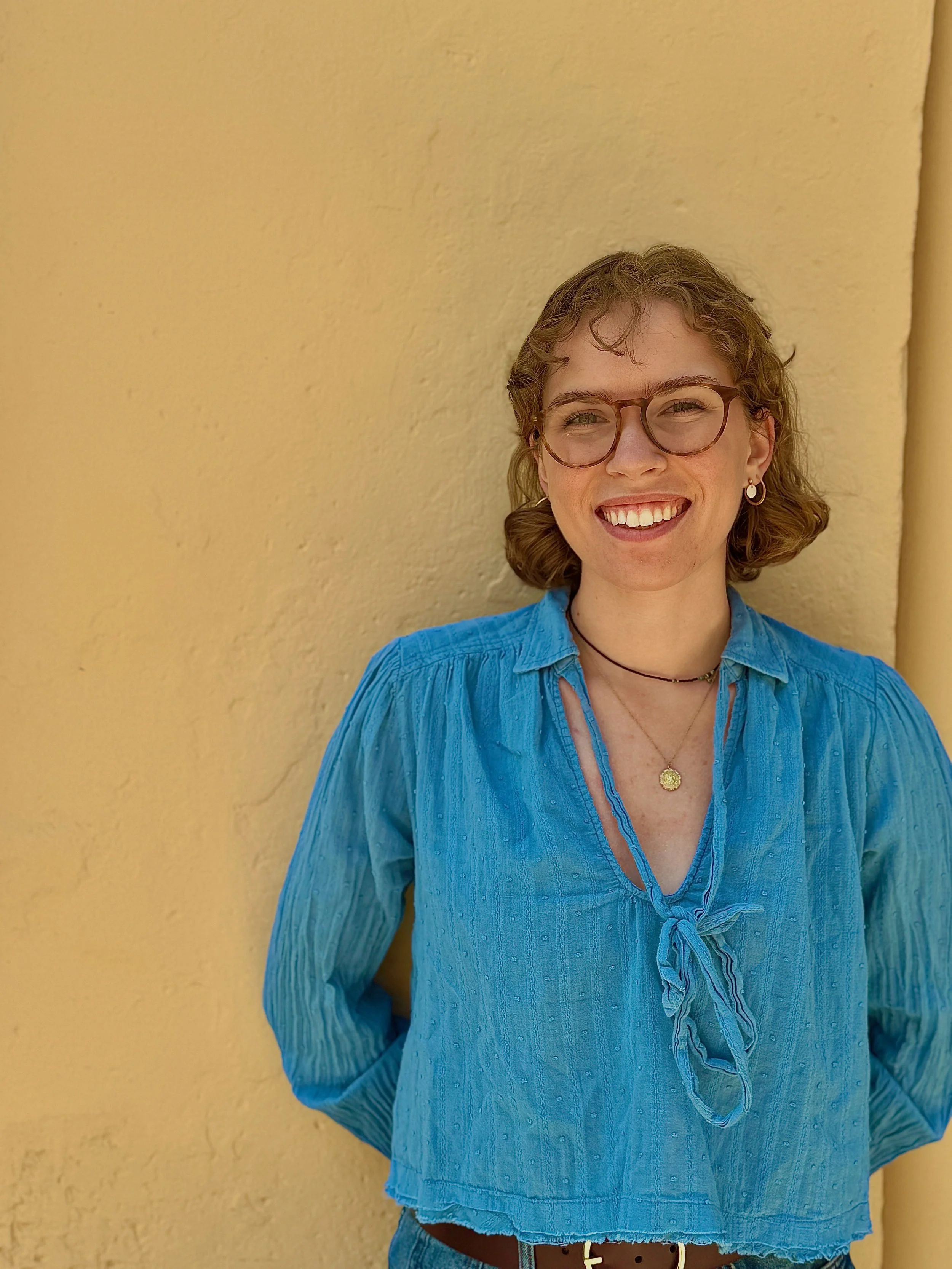 A young woman with curly hair, wearing glasses, earrings, a gold necklace, and a blue blouse, smiling against a yellow wall.