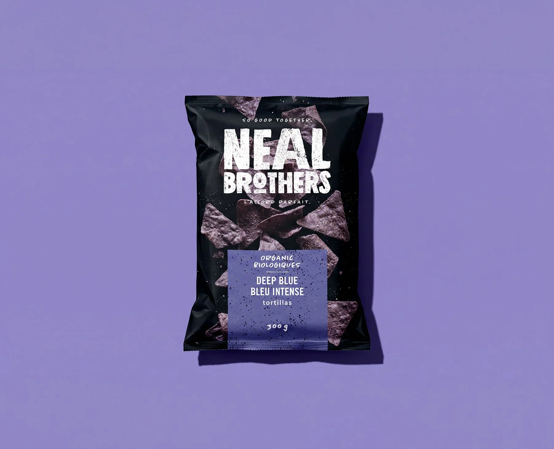 A black bag of Neal Brothers organic tortilla chips called Deep Blue, flavor described as bleu intense, weight 300 grams, on a purple background with the shadow cast to the right.