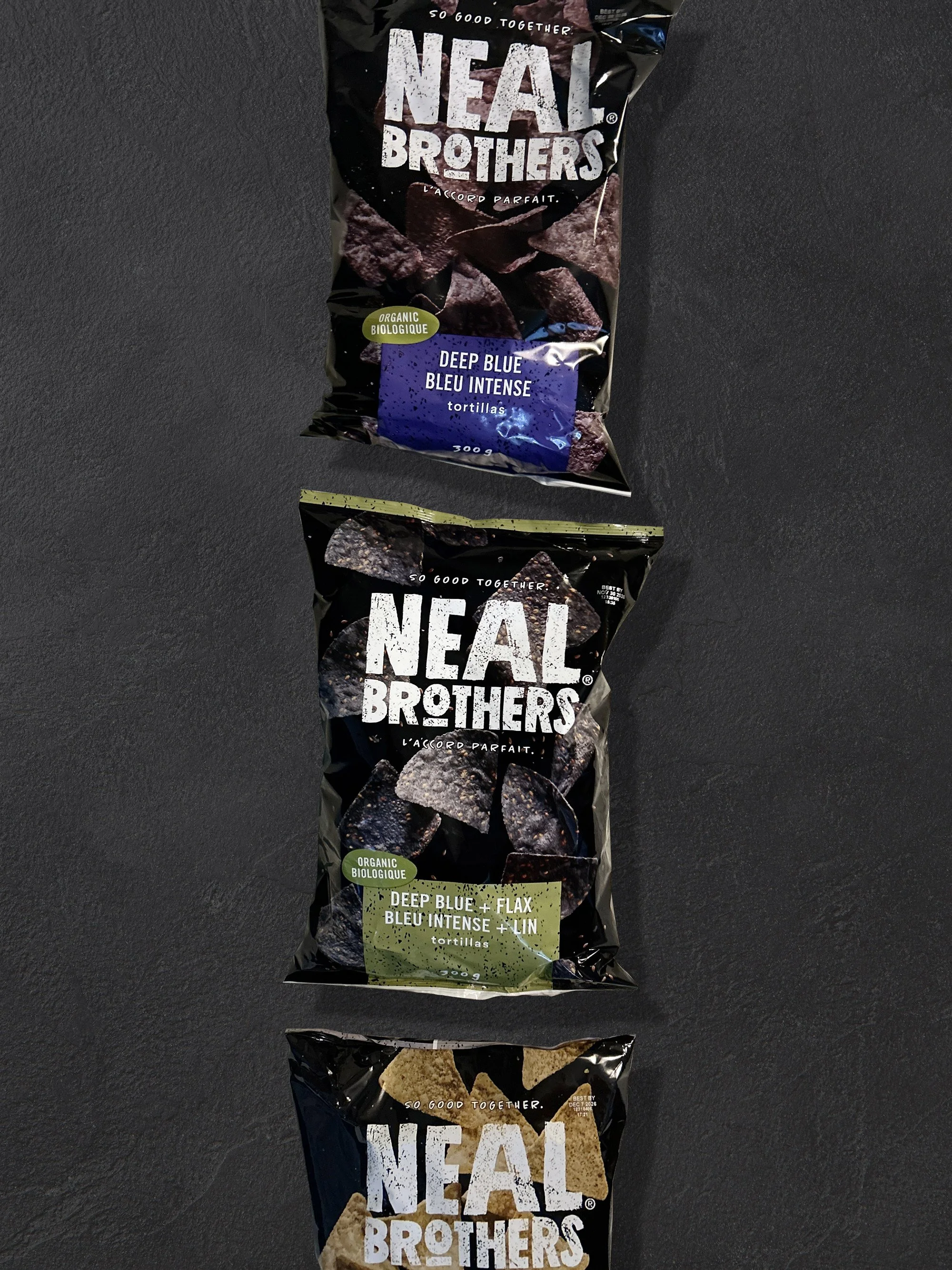 Three bags of Real Brothers organic tortilla chips stacked vertically on a black surface. The top bag is Deep Blue with blue-purple packaging, the middle bag is Deep Blue Flax with green packaging, and the bottom bag is tan-colored chips on black pac